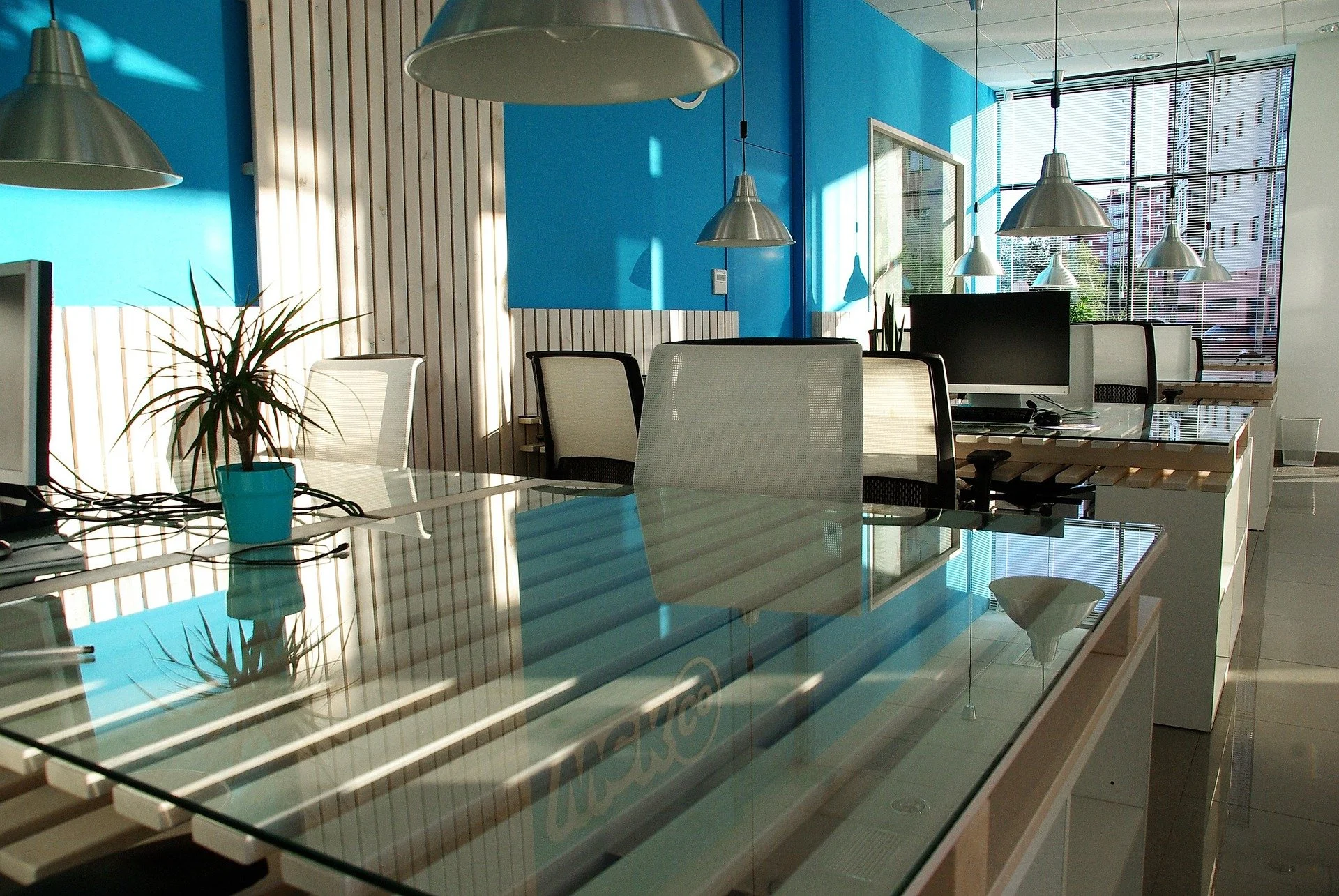 Modern office space with glass desks, white ergonomic chairs, blue accent wall, pendant lighting, and large windows with blinds.