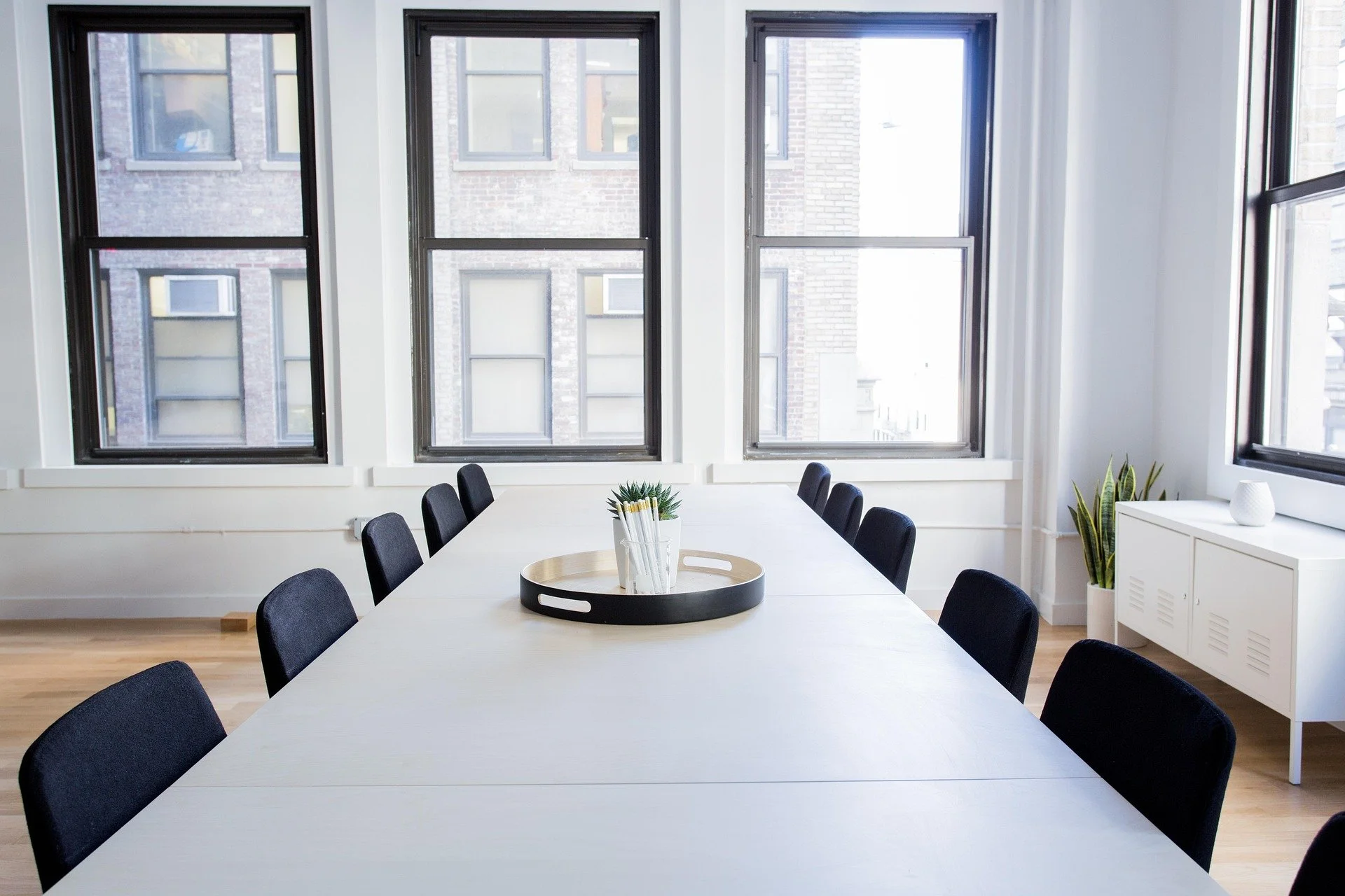 A bright modern conference room with a long white table, black chairs, large windows, a white sideboard with a potted plant, and a round tray with stationery in the center.