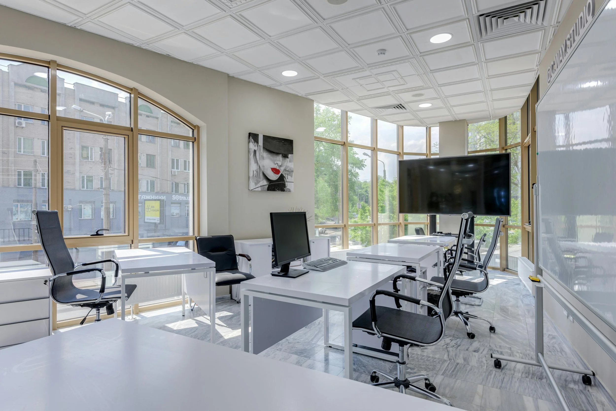 Modern conference room with white desks, black office chairs, large window with a city view, wall art, and a big TV screen.