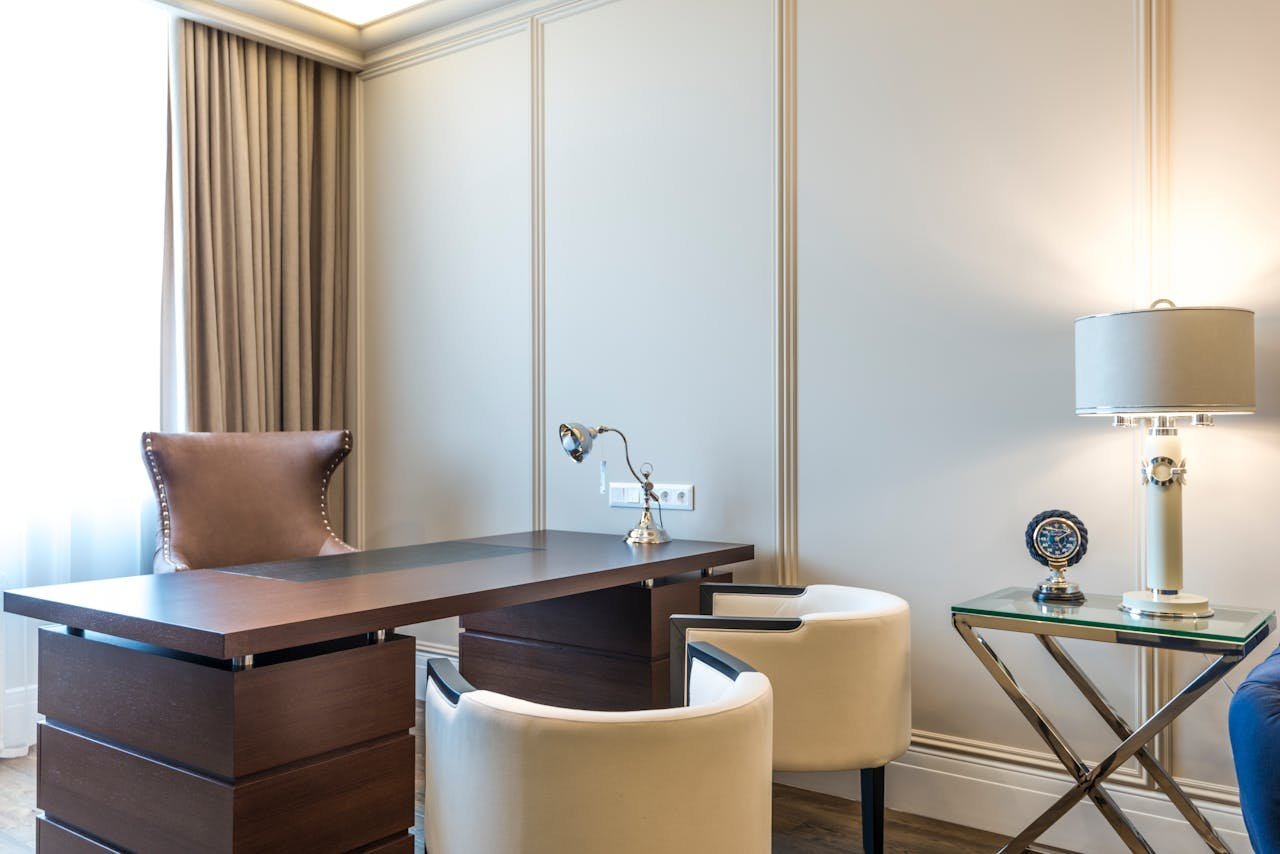 A modern office corner with a wooden desk, a leather chair with nailhead trim, two cream-colored chairs, a glass side table with a metallic base, a lamp, and a decorative clock.