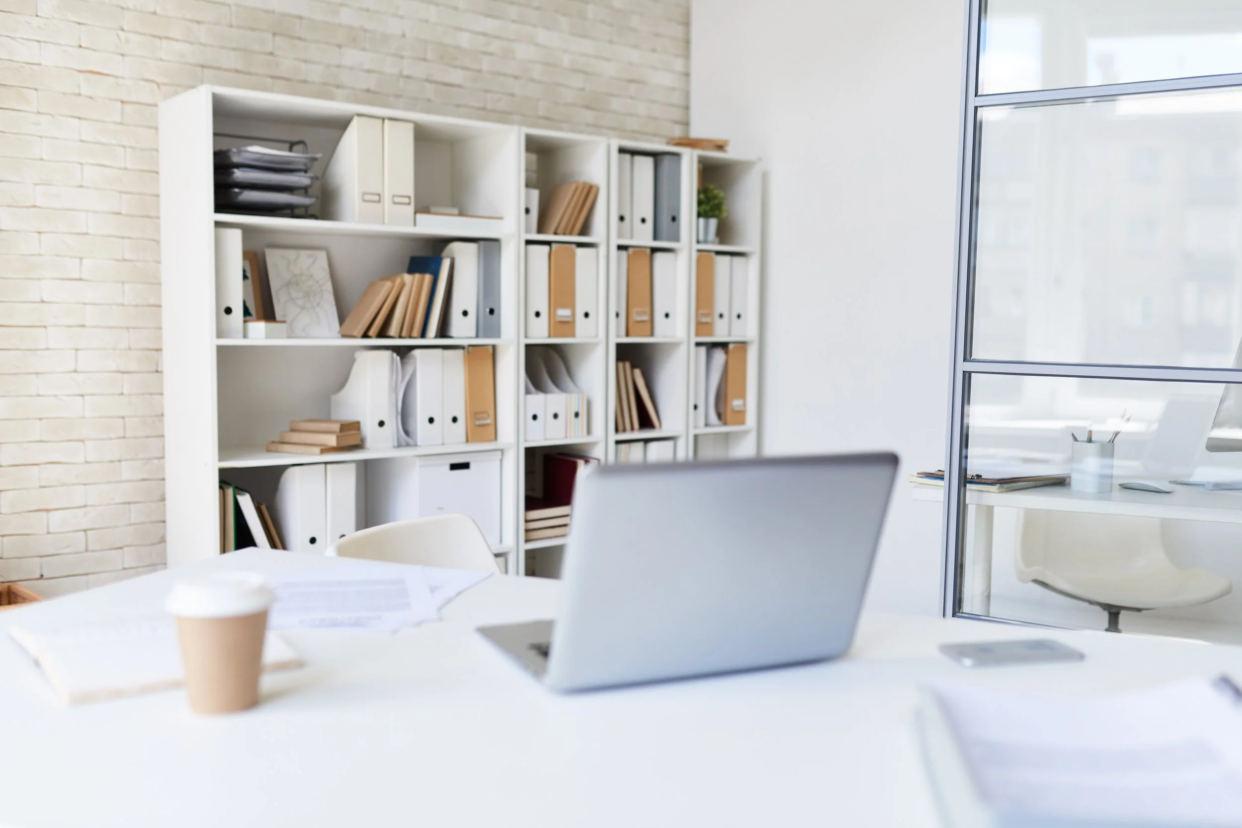Modern office with white walls, large window, white table with laptop, papers, and coffee cup, white bookshelf filled with binders and books, and a white chair.