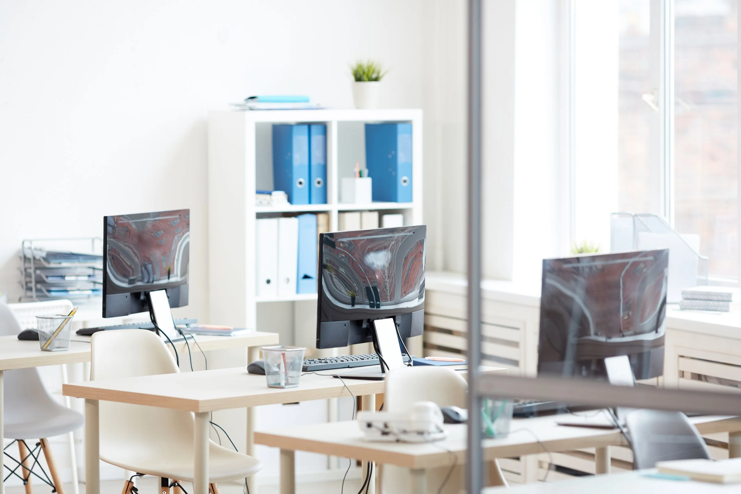 Office space with desks, computers, chairs, and a white bookshelf with blue binders, next to a window with natural light.