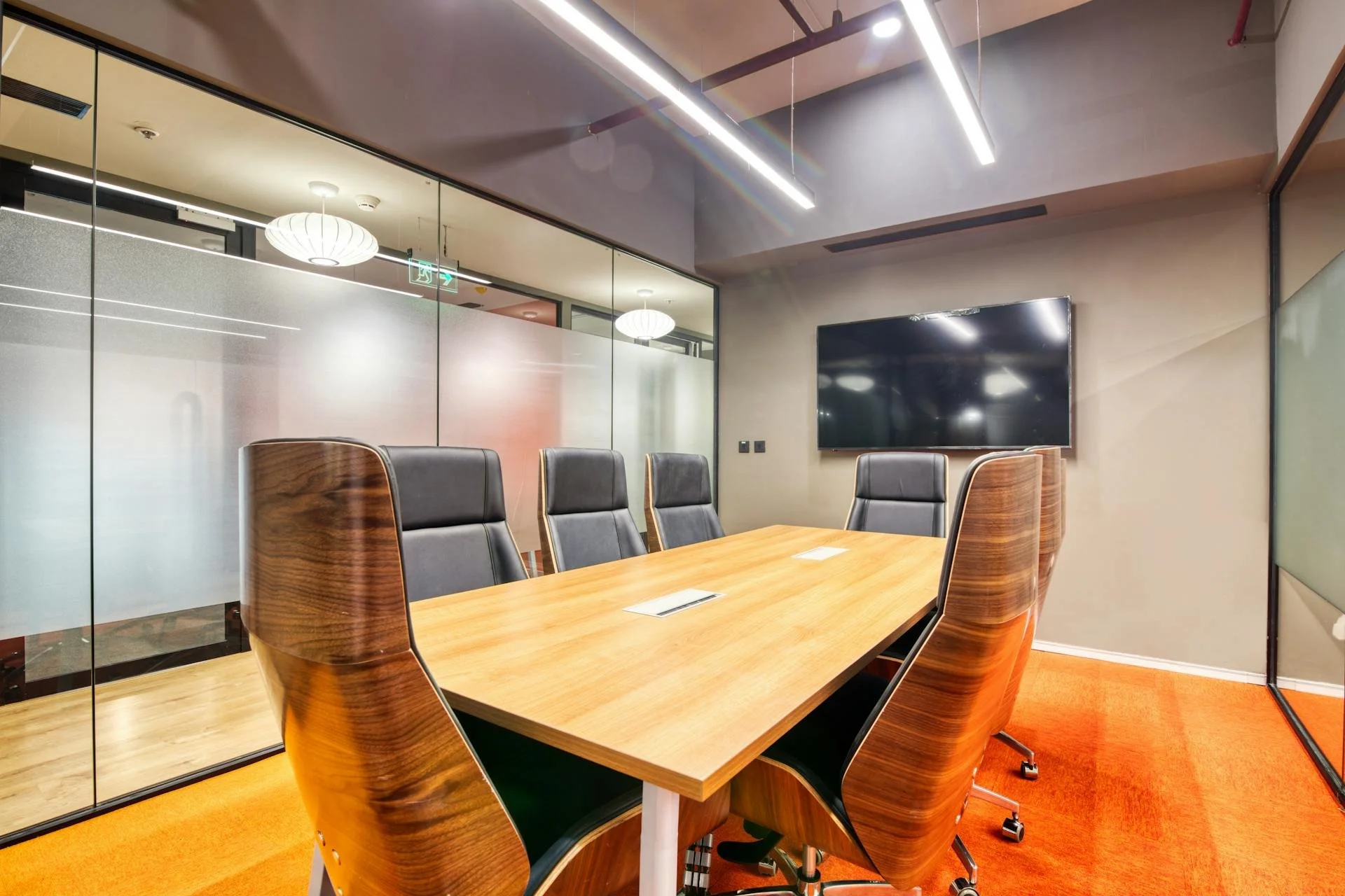 Modern conference room with a wooden table, six black leather chairs with wooden sides, a large wall-mounted TV, orange carpet, glass walls with frosted sections, and ceiling lighting.