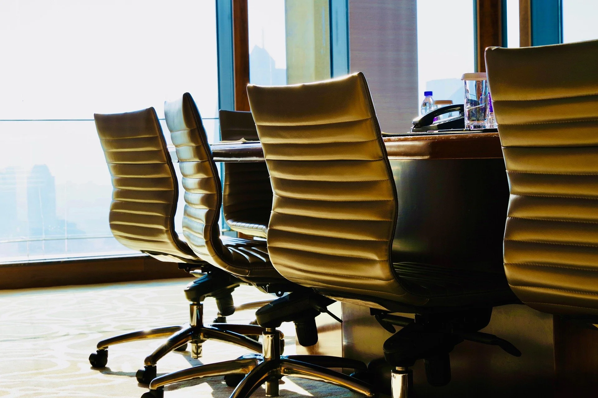 Empty conference room with leather chairs around a wooden table, large windows showing city skyline, and a bottle of water on the table.
