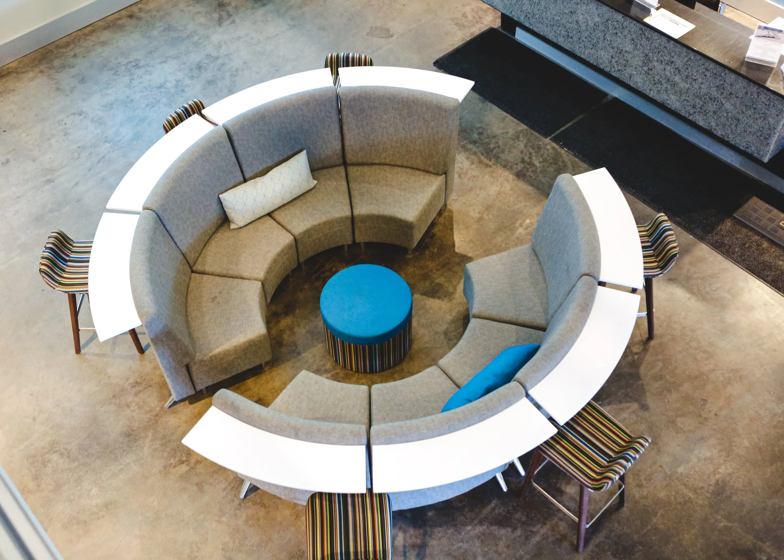 Top view of a circular lounge area with gray curved sofas, a small blue round ottoman, and three striped chairs around a marble-topped table.