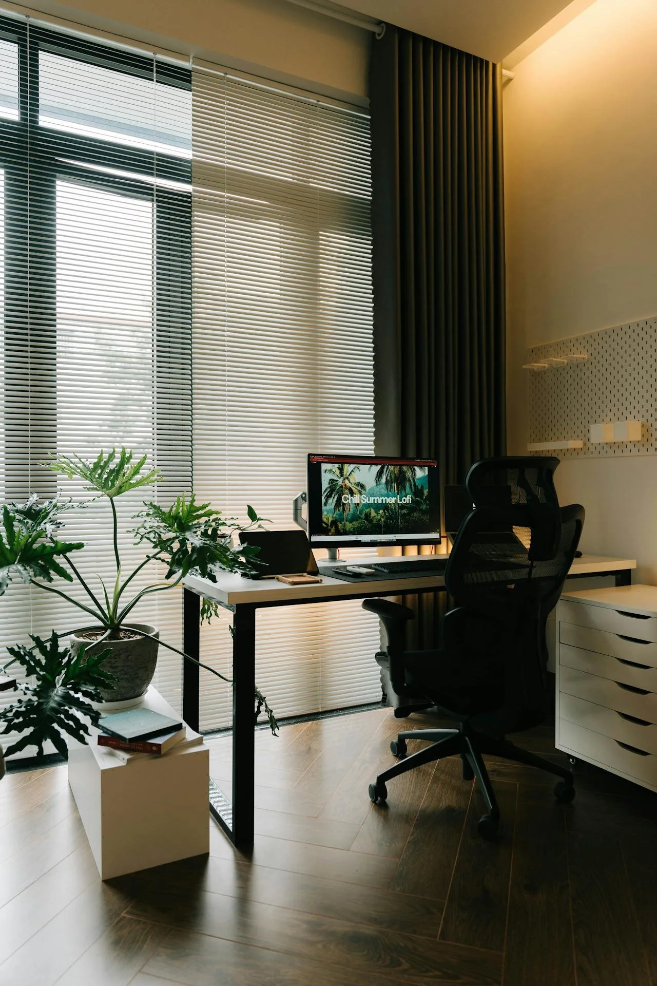 A modern home office with a desk, black ergonomic chair, potted plant, and computer displaying 'Chill Summer LoFi' against a window with closed blinds and vertical curtains.