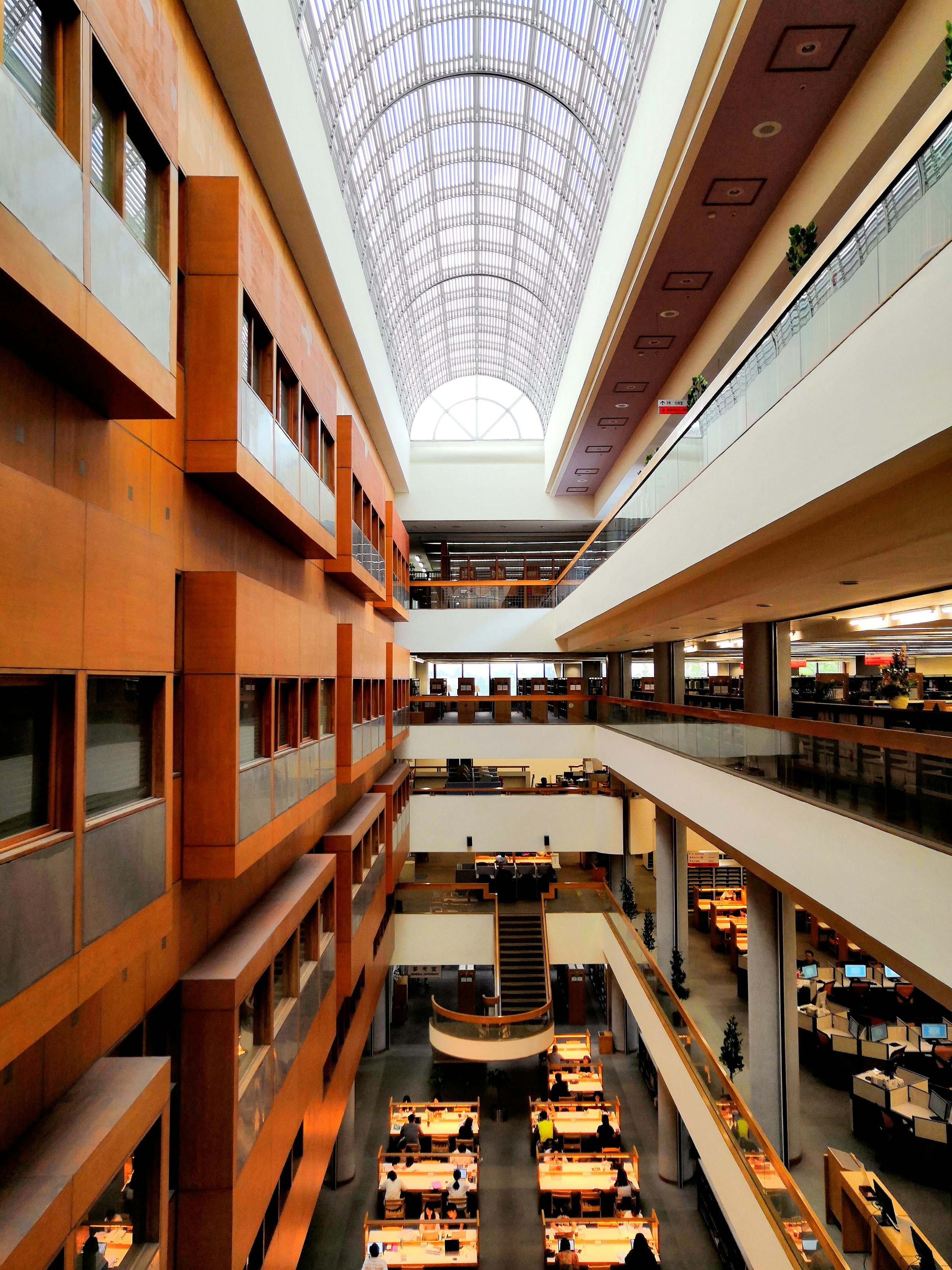 Multiple floors of a modern library or office building with wooden and white walls, glass railings, and a high glass ceiling allowing natural light to fill the space. People are working at desks.