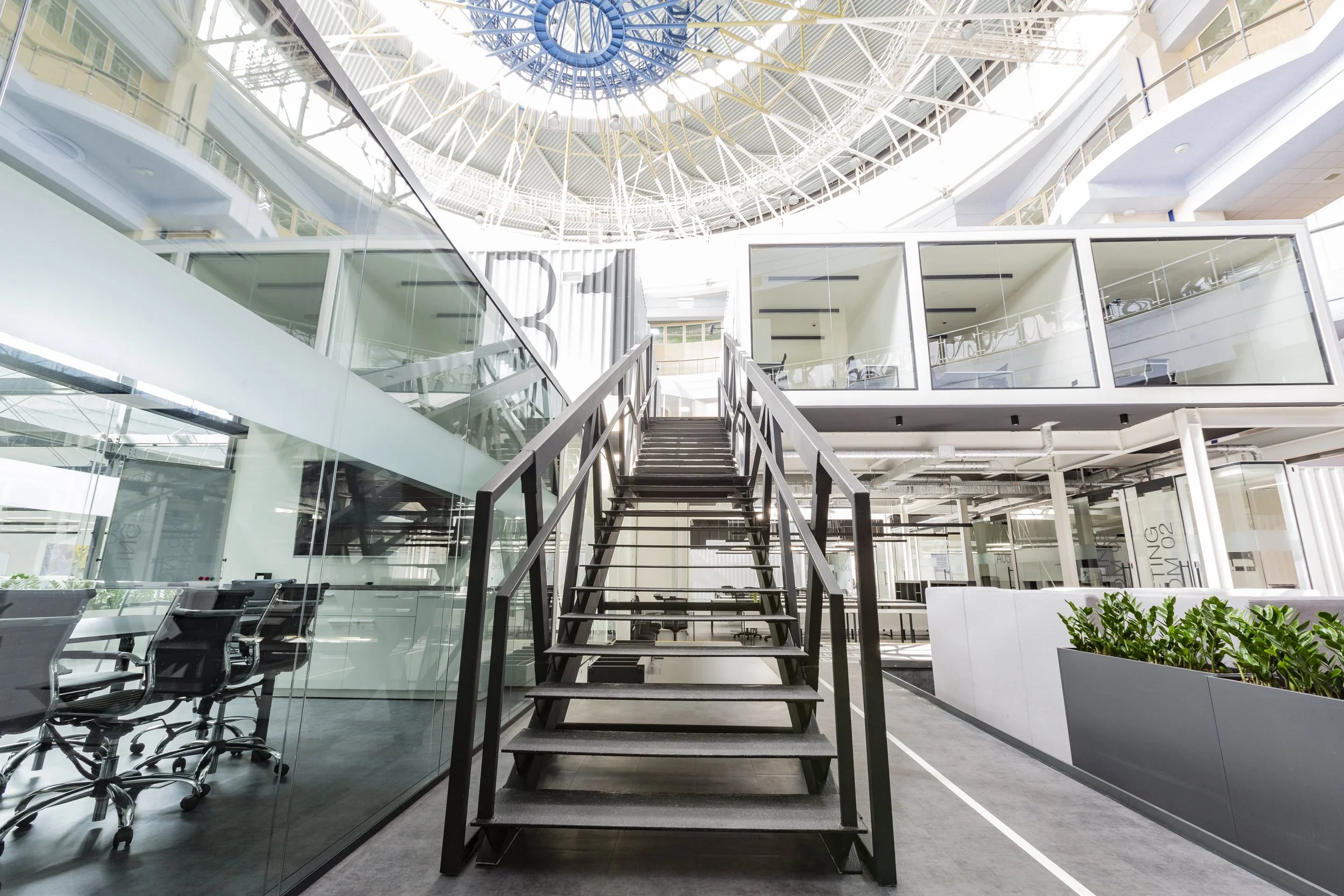 Modern office building interior with glass walls, a staircase at the center, and an open ceiling with a skylight.