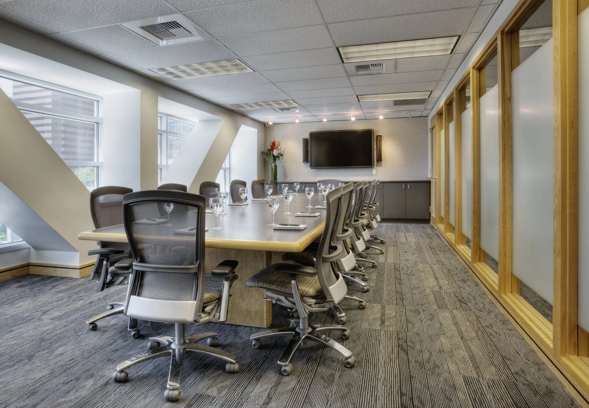 A modern conference room with a long oval table set with glasses and notepads, surrounded by office chairs, with a large flat-screen TV, window lighting, and decorative wall panels.