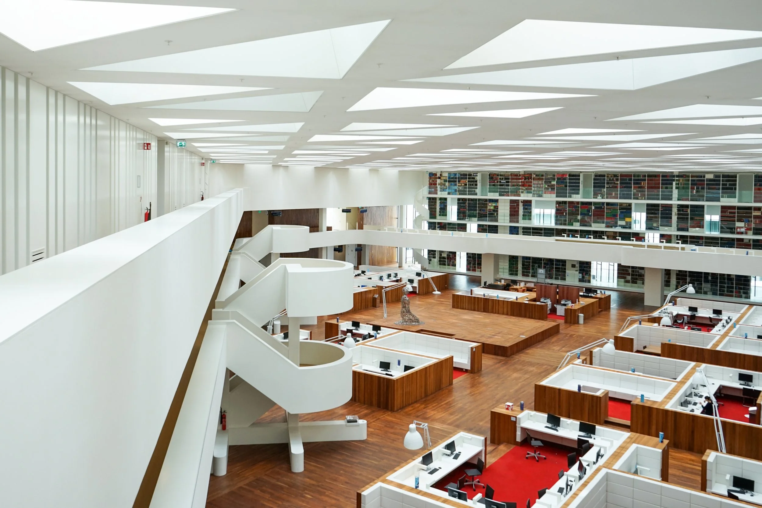 Spacious modern library interior with wooden floors, white walls, contemporary art sculpture, glass bookshelves, and multiple workstations with computers and office chairs.