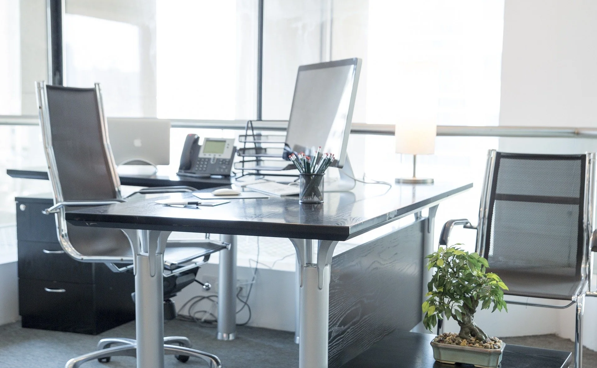 Empty modern office with a desk, two chairs, a computer monitor, a landline phone, and a desk lamp, a potted bonsai plant next to the desk