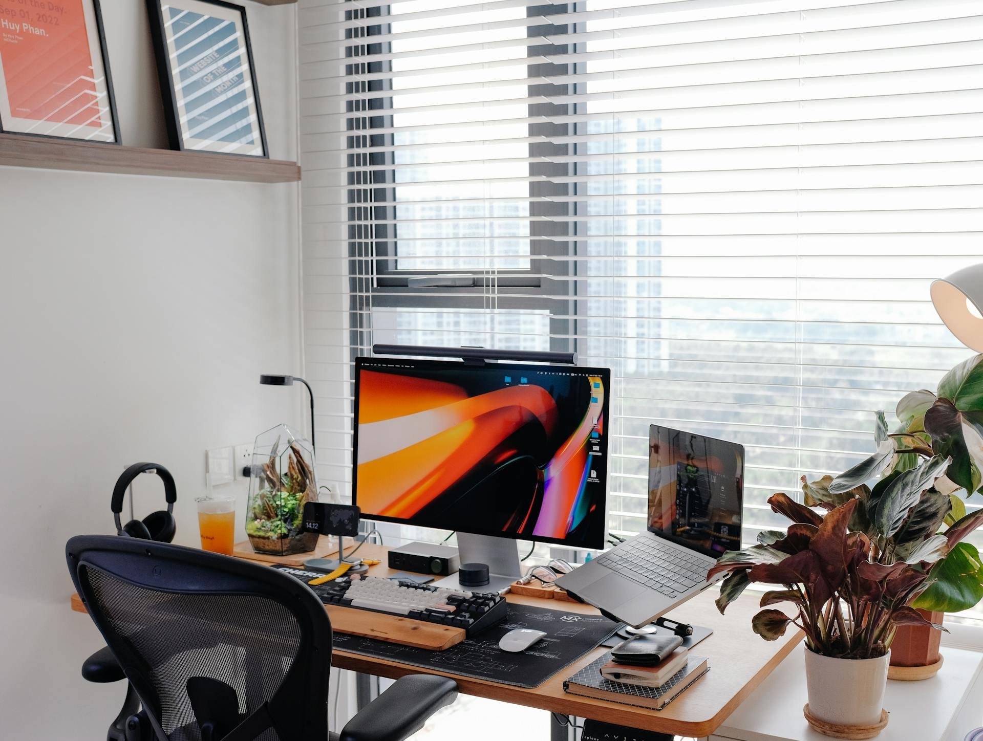 Modern home office desk with computer monitor, laptop, books, plants, and headphones near large windows with blinds