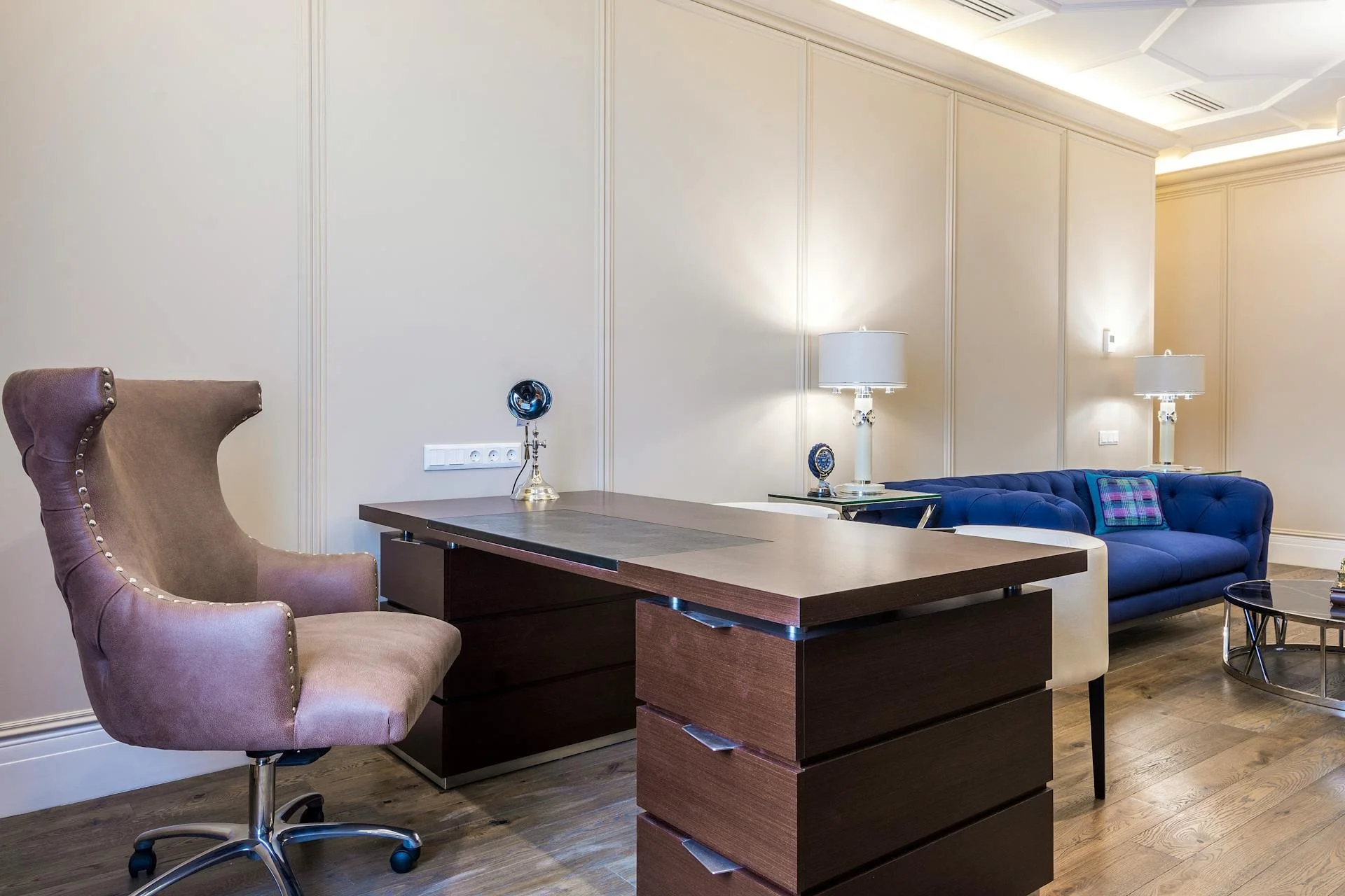 Modern office space featuring a beige high-back swivel chair, a dark wooden desk with multiple drawers, two matching table lamps on side tables, and a blue tufted sofa with a plaid cushion, set in a room with cream-colored walls and wood flooring.