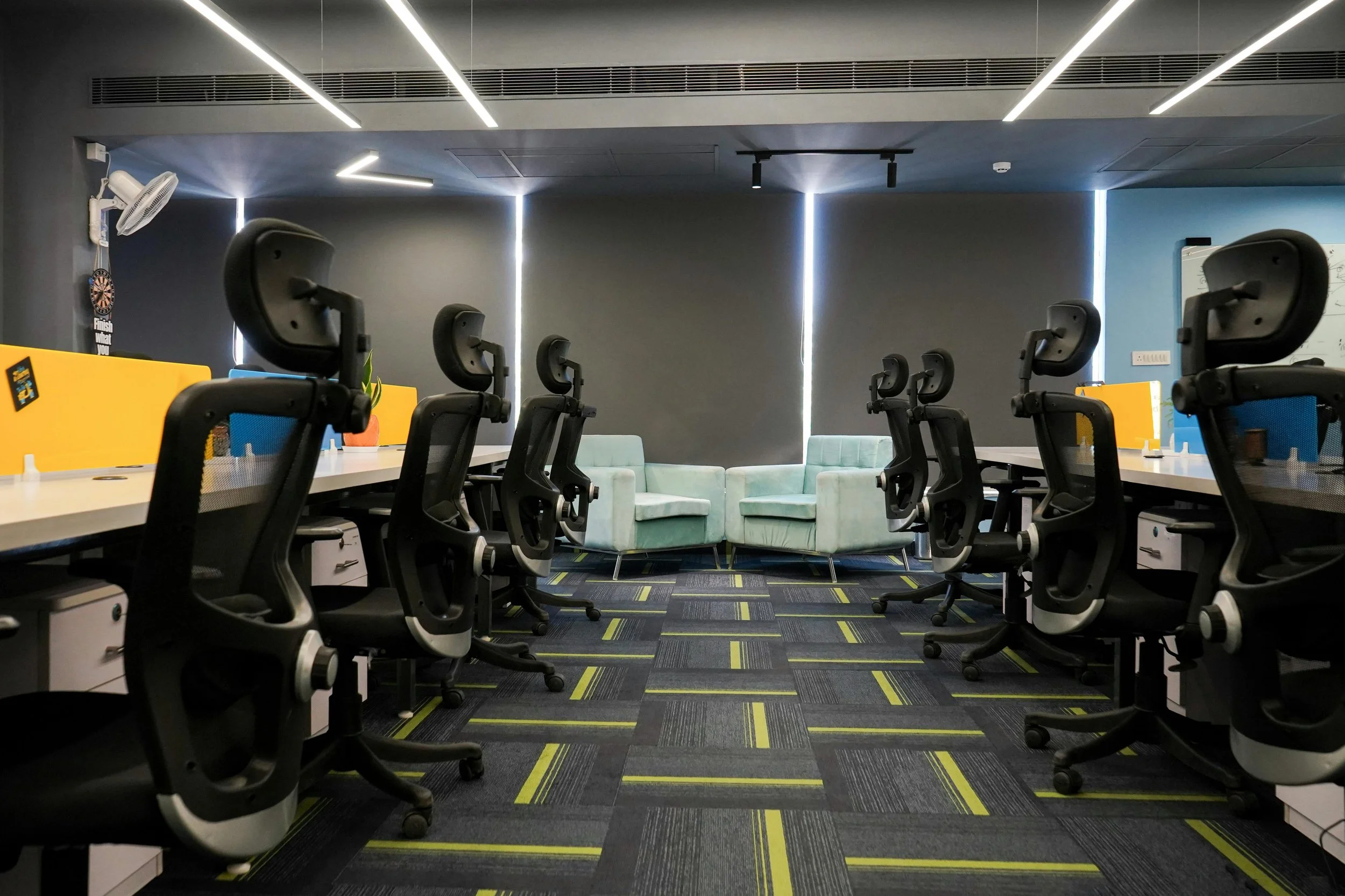 An empty modern office with rows of black ergonomic office chairs facing a long desk, and a lounge area with light green armchairs in the background. The office has dark walls, striped carpet, and vertical light strips on the wall.