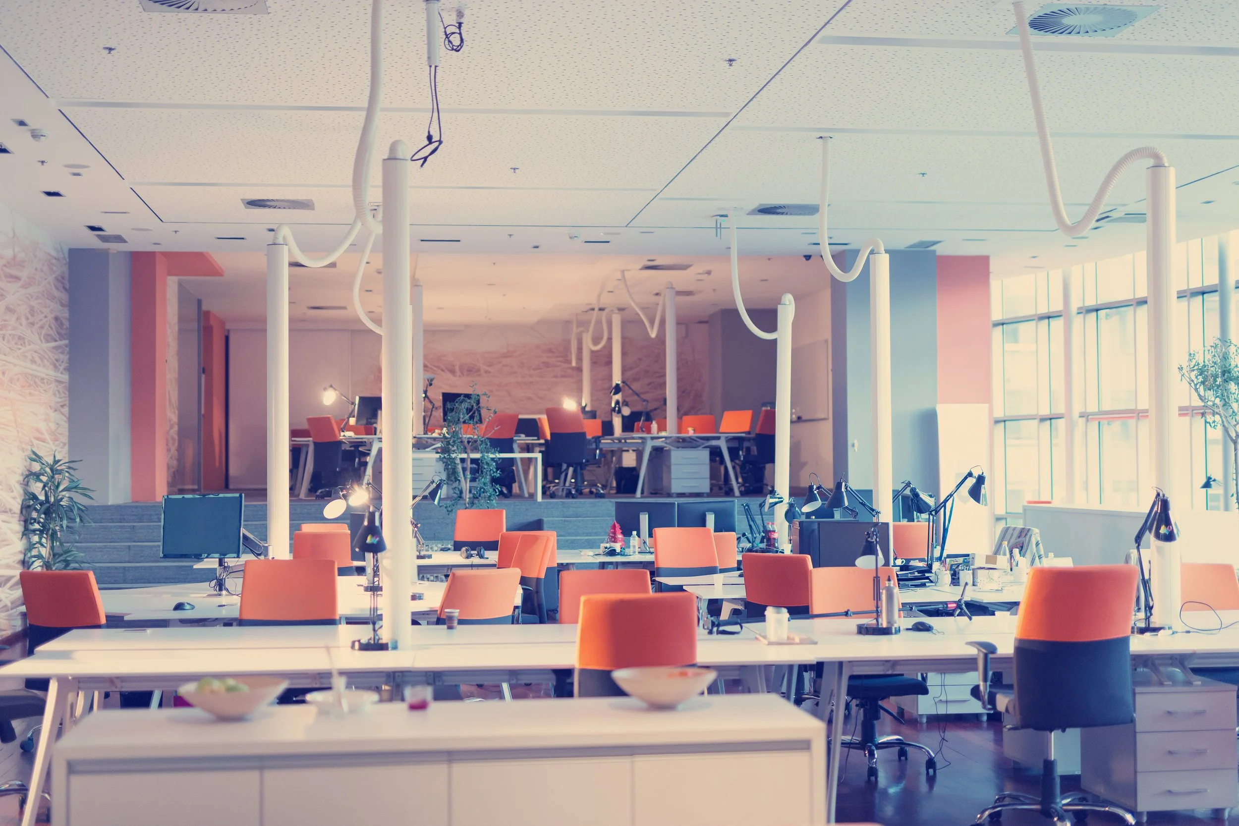 Empty modern office with orange chairs, desks, computer monitors, and standing lamps, near large windows letting in natural light.
