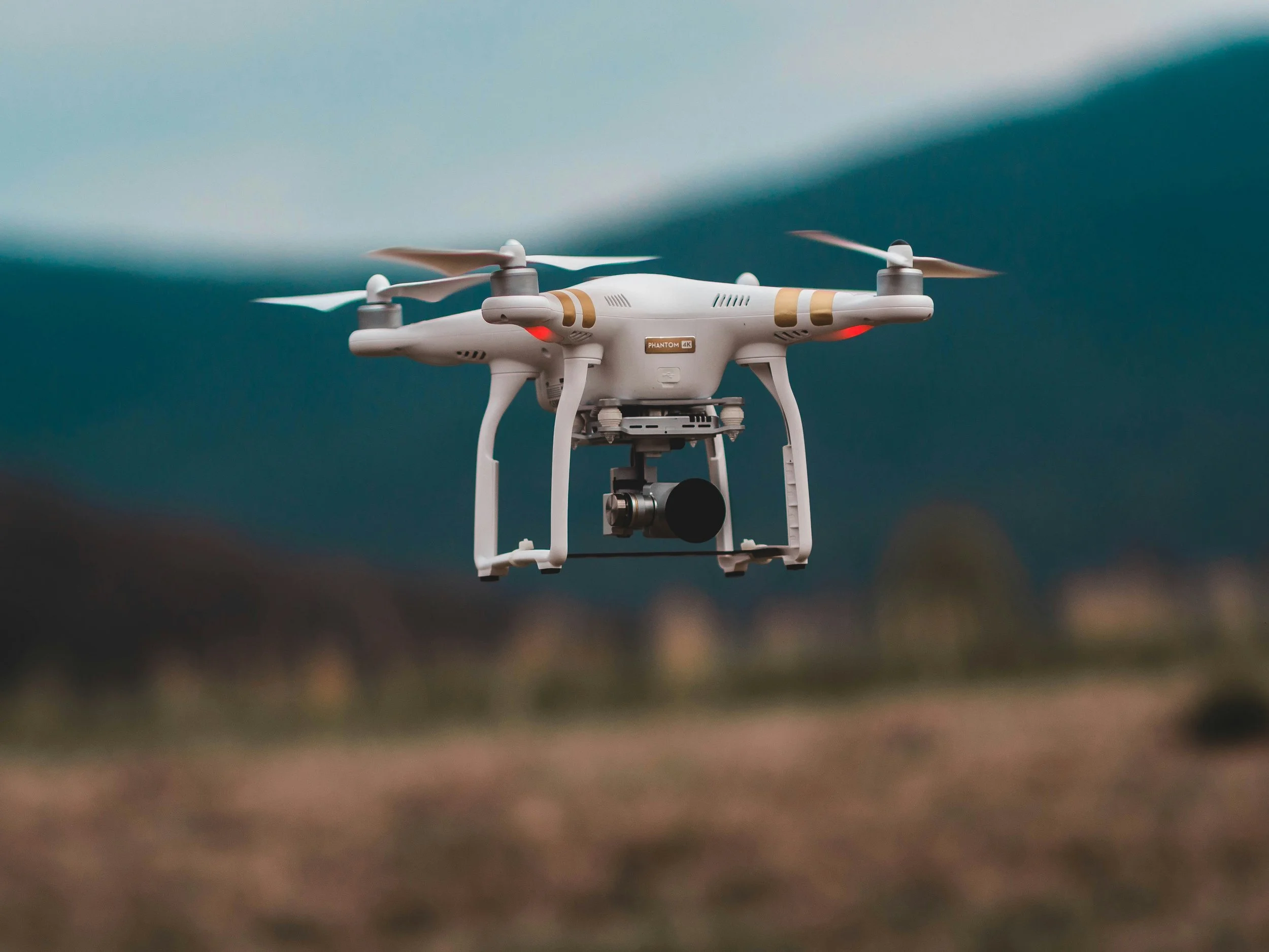 A white quadcopter drone flying over a blurred landscape with mountains and fields in the background.