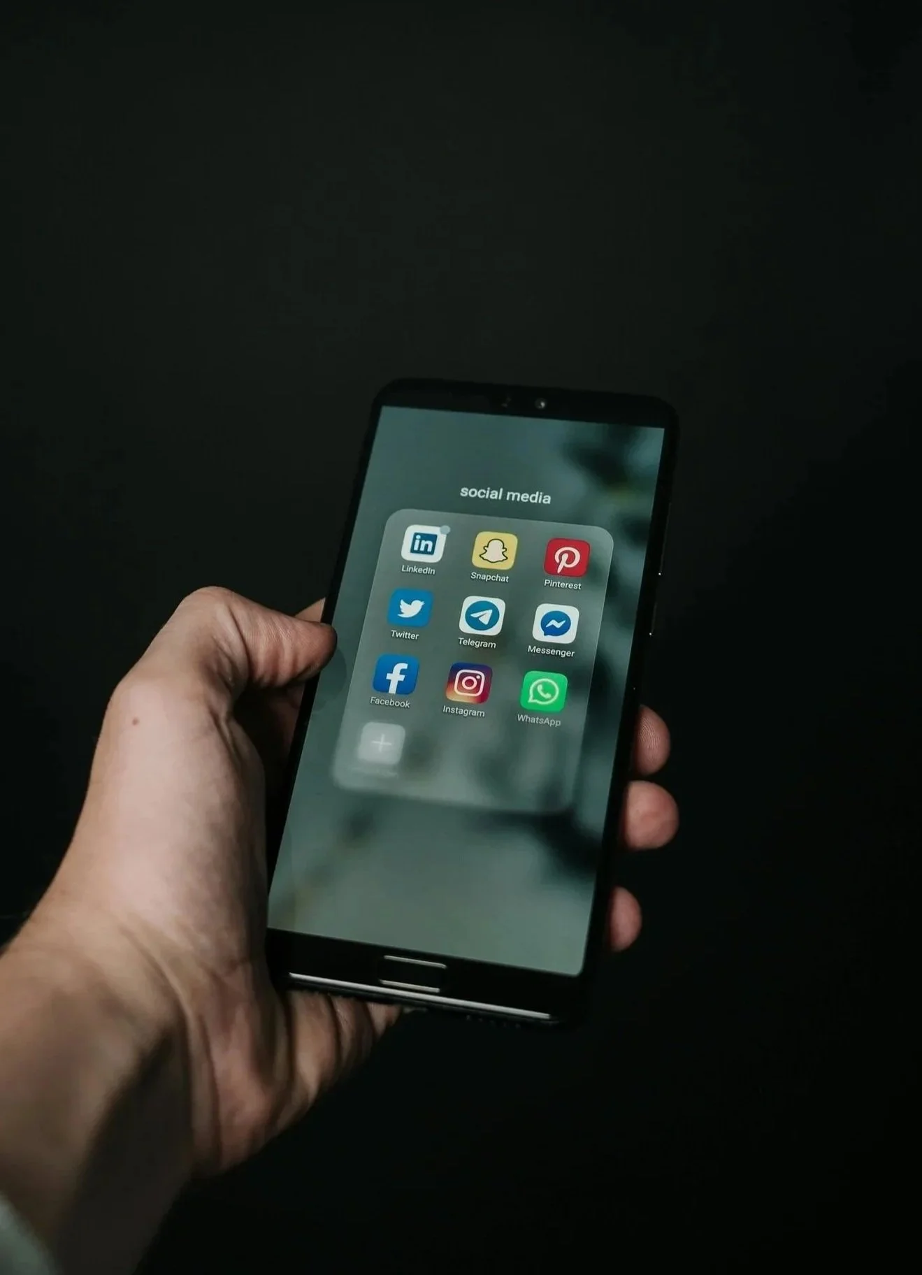 Hand holding smartphone displaying social media app icons in a folder labeled 'social media' against a dark background.