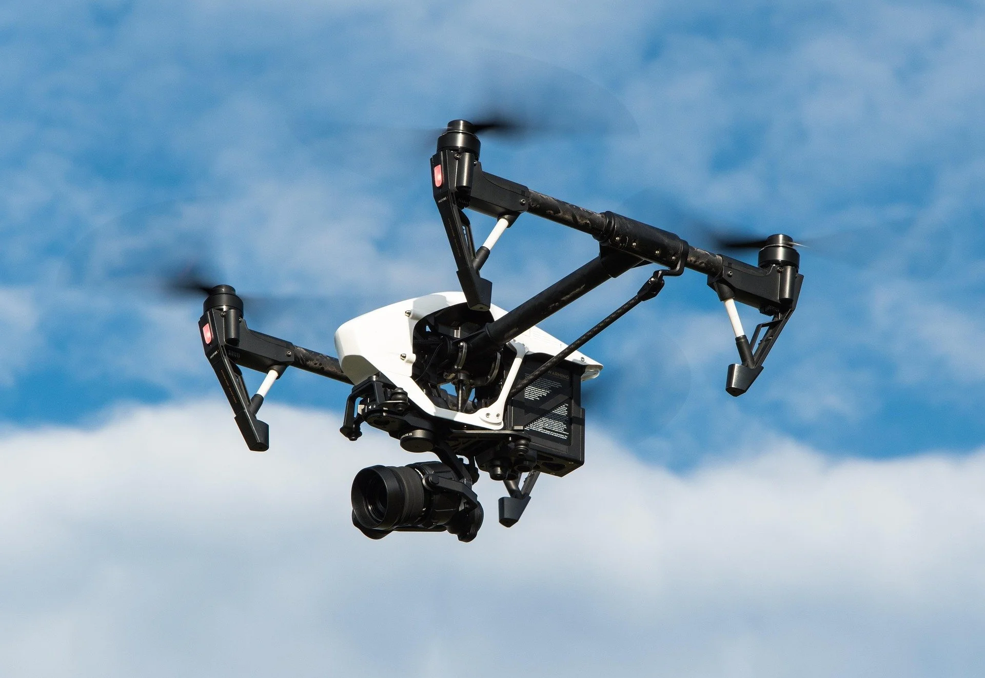 A drone with a camera mounted on underside, flying against a background of partly cloudy sky.