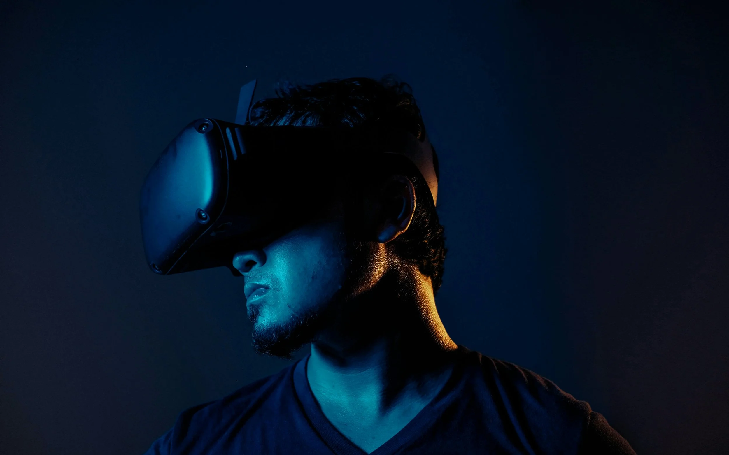 A man with dark hair and a beard wearing a virtual reality headset against a dark background with blue and orange lighting.