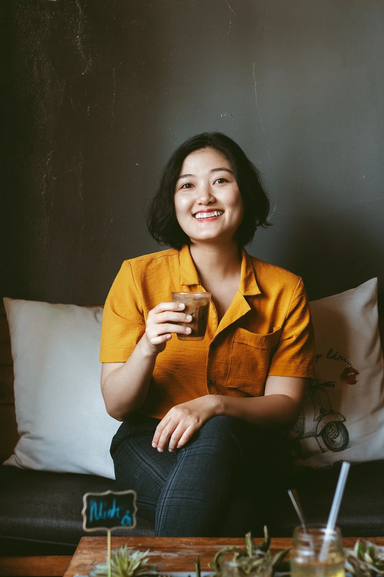 A woman with short black hair smiling while holding a glass of iced coffee, sitting on a couch with white pillows, wearing an amber-colored shirt, against a dark gray wall.