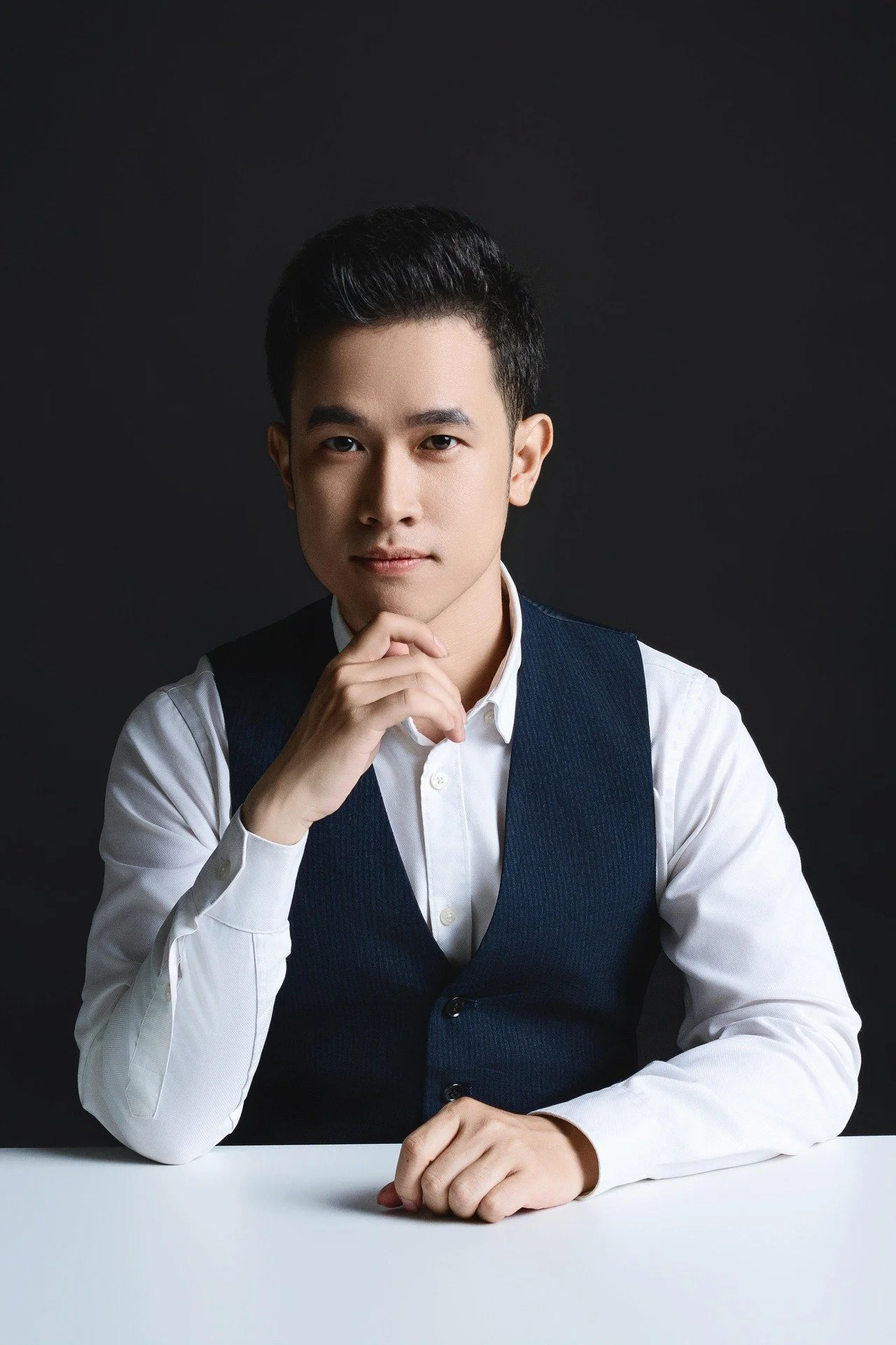 Portrait of a young man with black hair, wearing a white shirt and a dark vest, sitting at a white table against a dark background, with his chin resting on his hand.