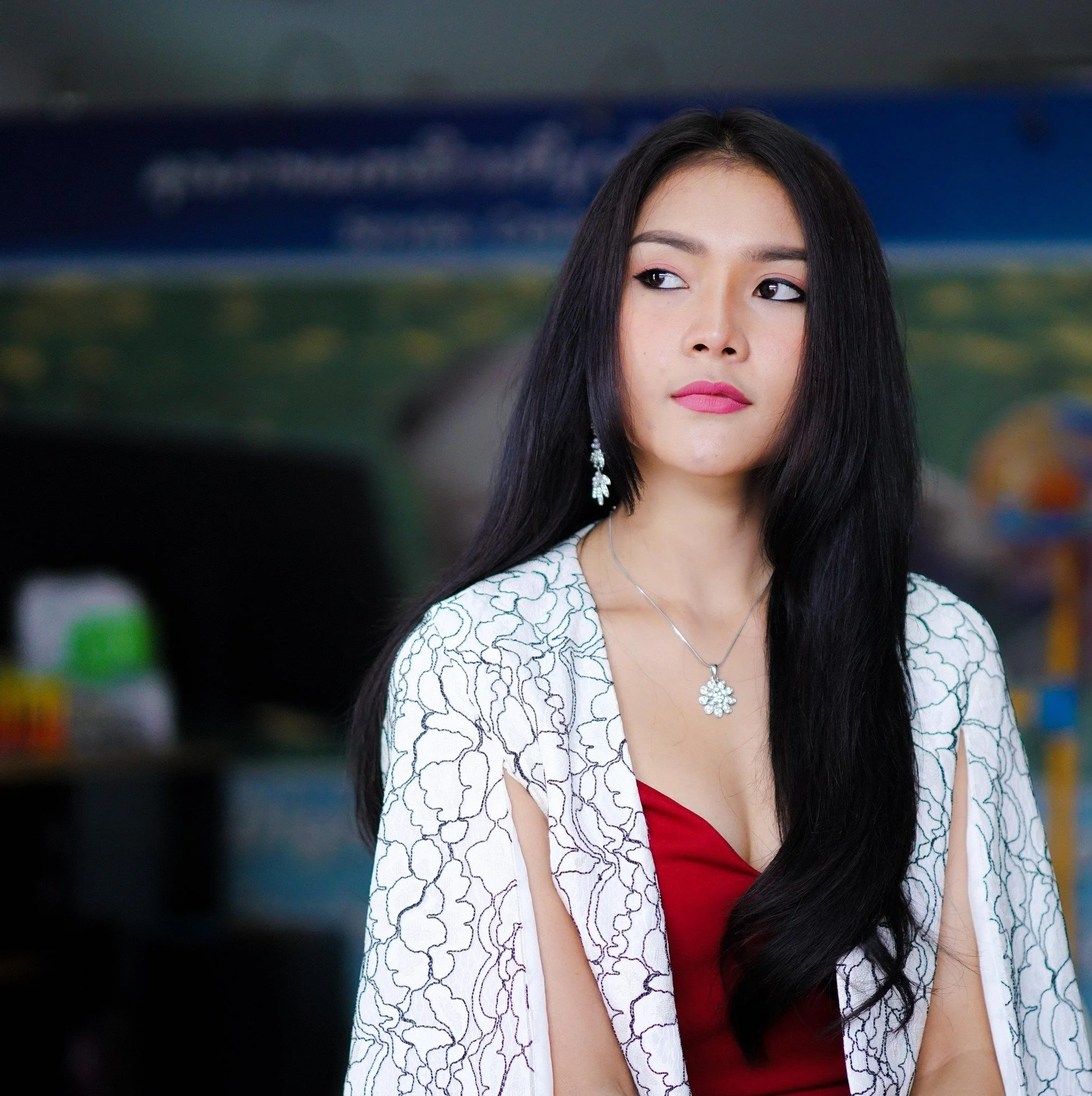 A woman with long black hair wearing a white lace cover-up, red dress, and jewelry, standing indoors.