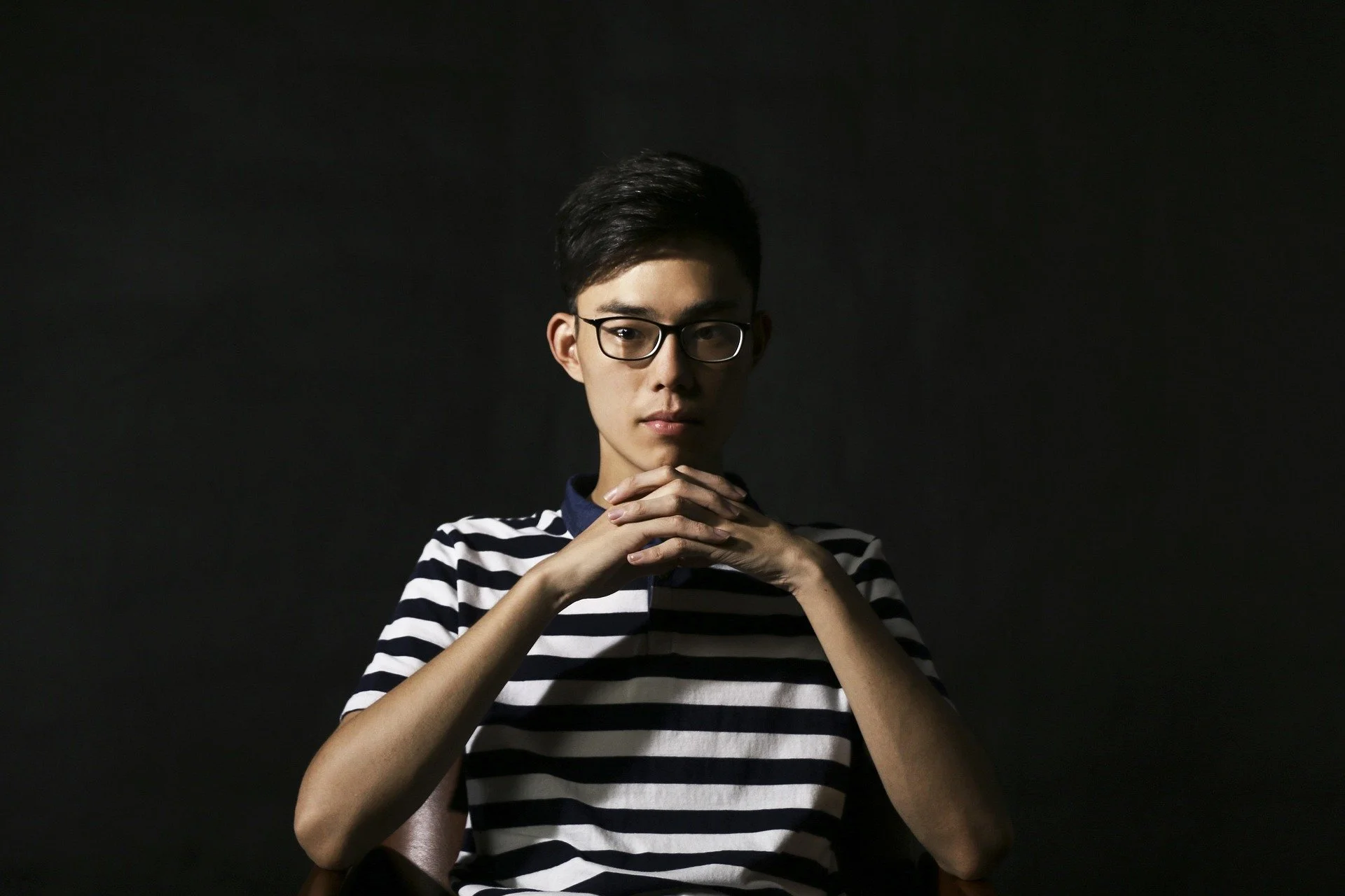 Young person with short black hair and glasses, wearing a black and white striped shirt, sitting in front of a dark background, looking at the camera with hands clasped.