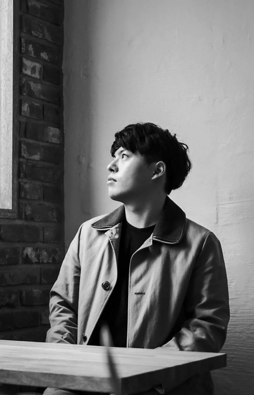 Black and white photo of a young man sitting at a table, looking to his left, wearing a jacket, with a brick wall and plain wall in the background.