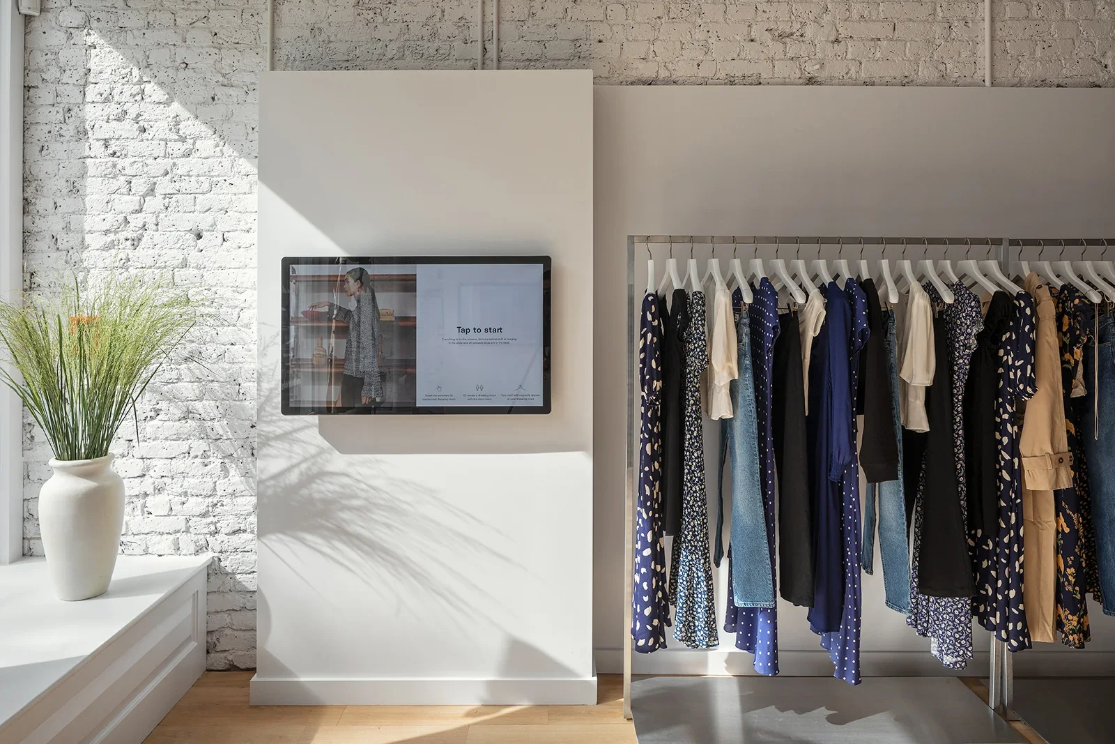Clothing store with a rack of various dresses and tops in shades of blue, white, black, and beige. A flat screen monitor displaying a woman in a store, with text 'Tap to start'. A white door with a potted plant is on the left side of the image.