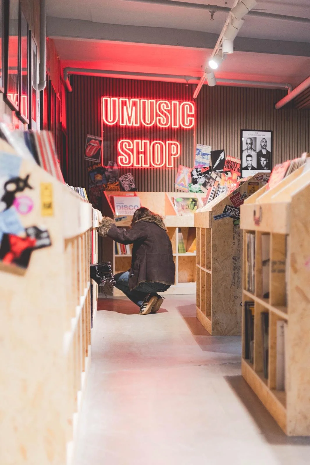 Person browsing vinyl records in a music shop with a neon 'U MUSIC SHOP' sign on the wall.