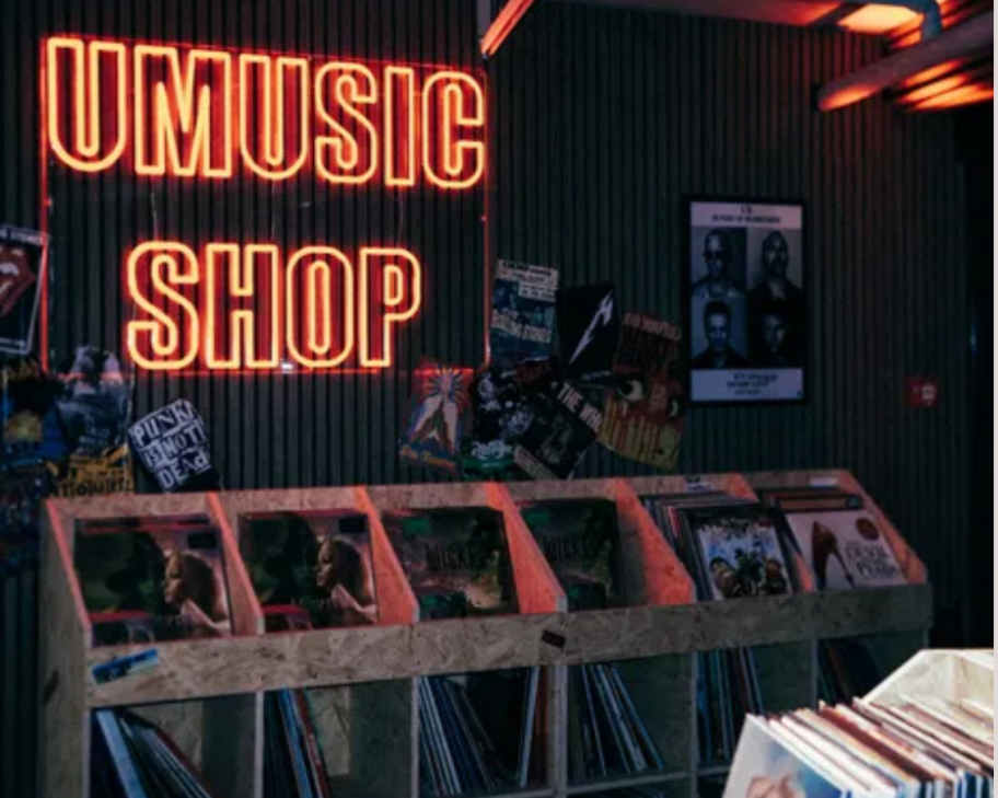 Red neon sign that reads 'UMusic Shop' hangs above a display of vinyl records and magazines, with posters on the wall behind.