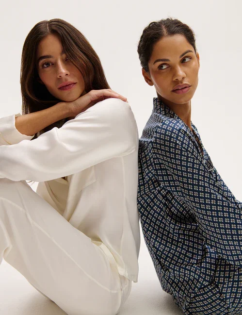 Two women sitting with their backs against each other, looking in opposite directions, against a plain light background.