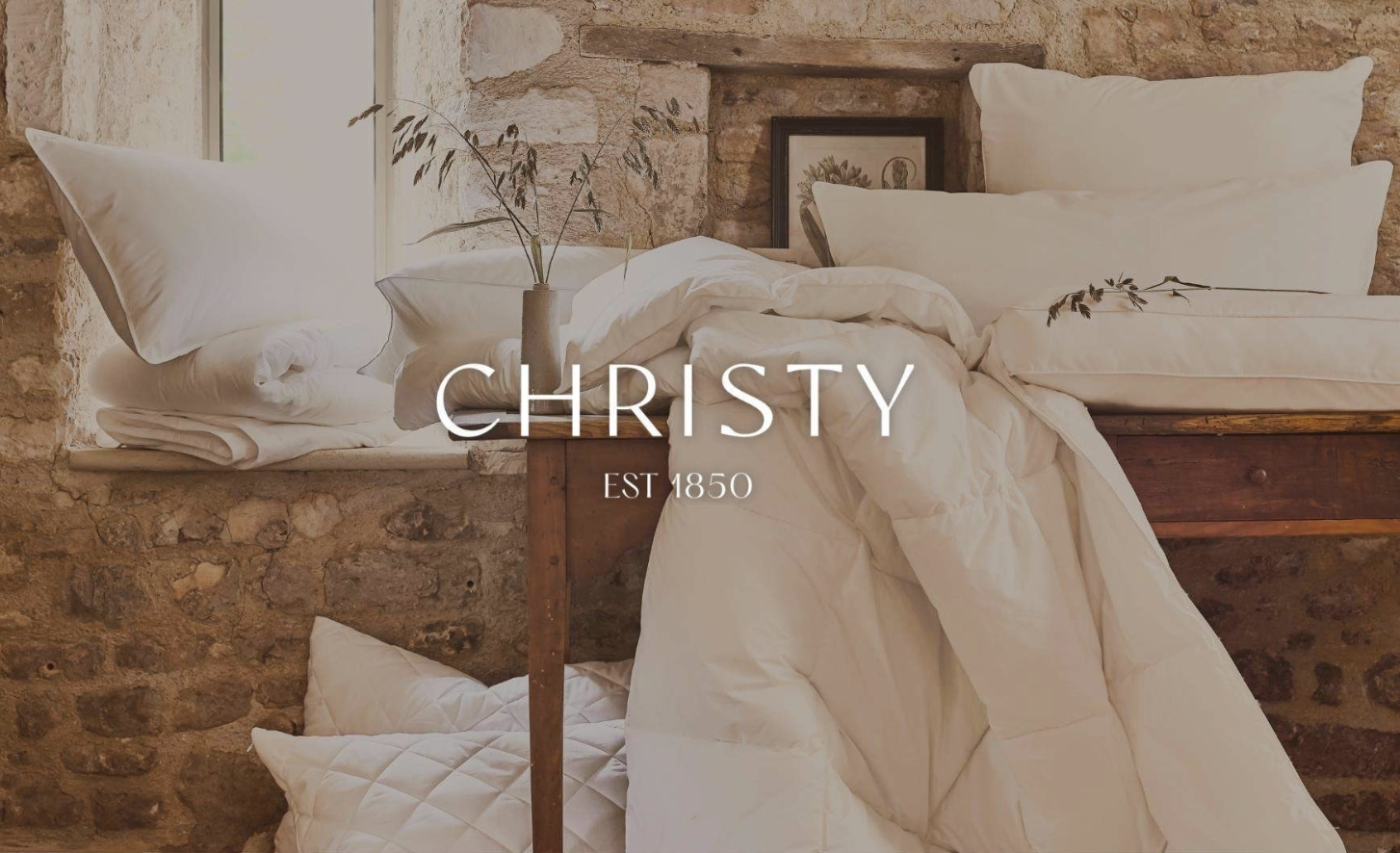 A rustic bedroom with a stone wall, showing a bed with white bedding, pillows, and a wooden side table with a vase of dried grass, and a framed botanical print in the background. The logo 'CHRISTY" in white.