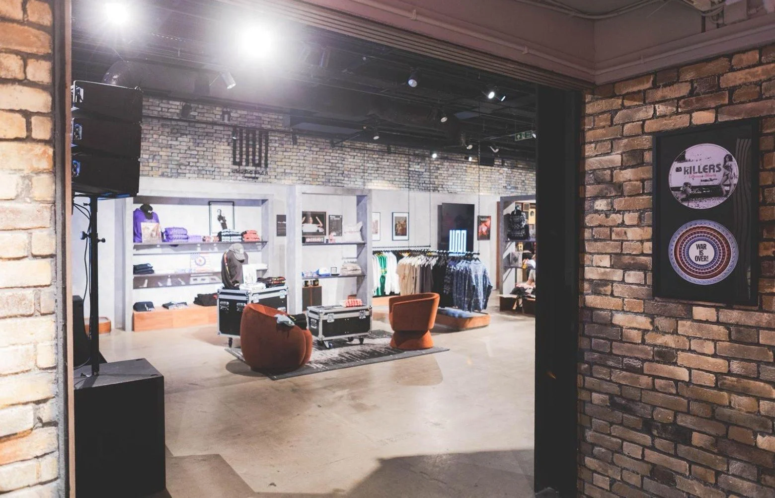 View of a retail clothing store entrance with exposed brick walls, showcasing shelves with folded clothes, shirts on hangers, artwork on the wall, and two orange chairs in the center.