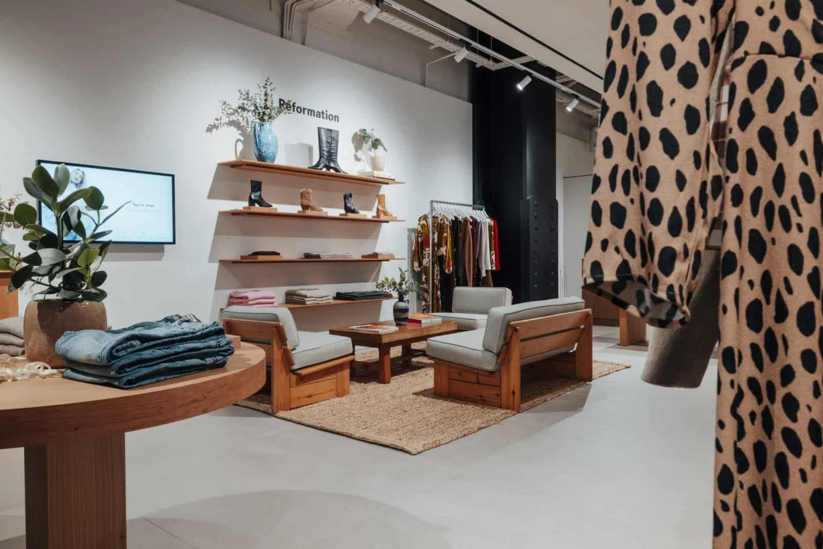 Interior of a boutique clothing store with wooden furniture, shelves displaying shoes and accessories, a clothing rack with colorful garments, and seating area with white cushioned chairs. A person wearing a leopard-print coat is partially visible on the right.