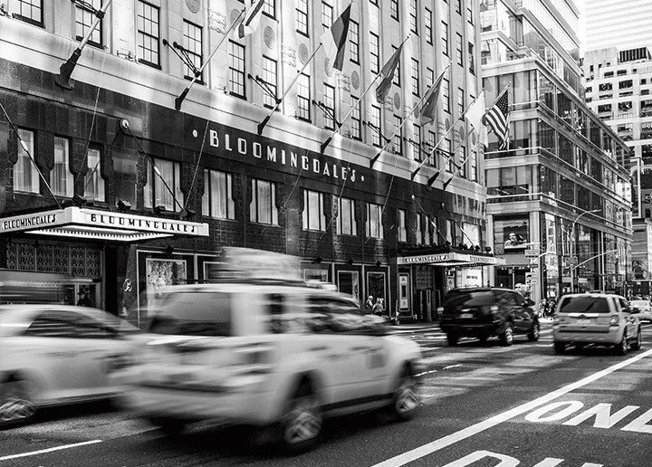 Black and white photo of Bloomingdale's department store storefront on a busy city street with cars moving, flags hanging above, and urban buildings.