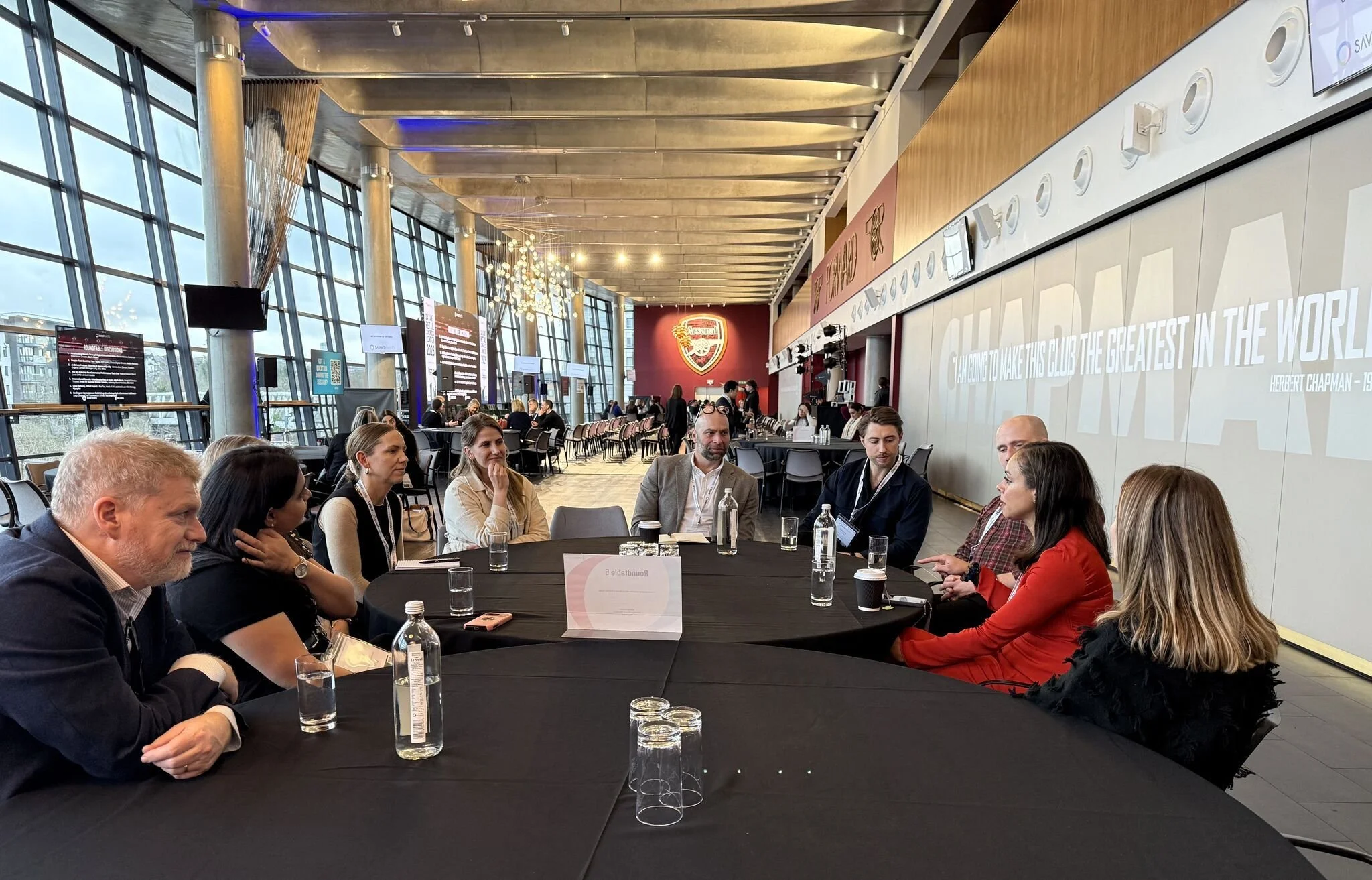 A group of people sitting at a round table during a conference or meeting in a modern, spacious venue with large windows, a red Arsenal logo on the wall, and a large quote on the wall reading 'Striving to make this club the greatest in the world'.