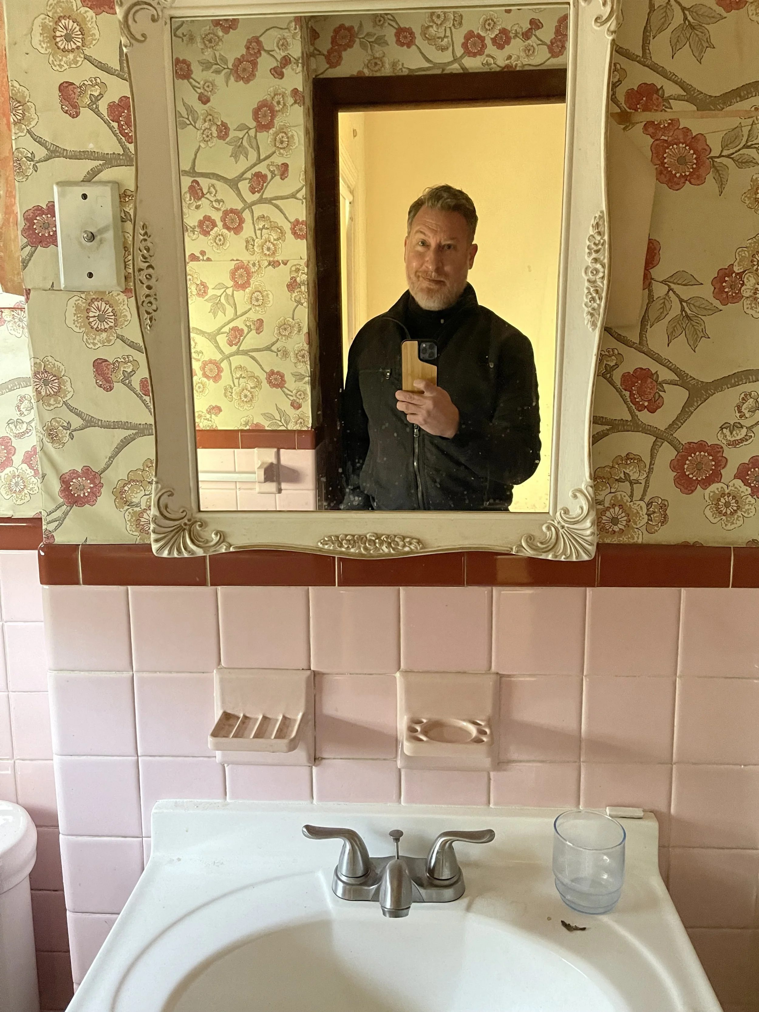 A bathroom sink with a mirror above it, reflecting a person taking a selfie. The wall behind has floral wallpaper, and there are pink tiles above the sink. A glass is on the right side of the sink, and there are small soap dishes or holders mounted on the wall.