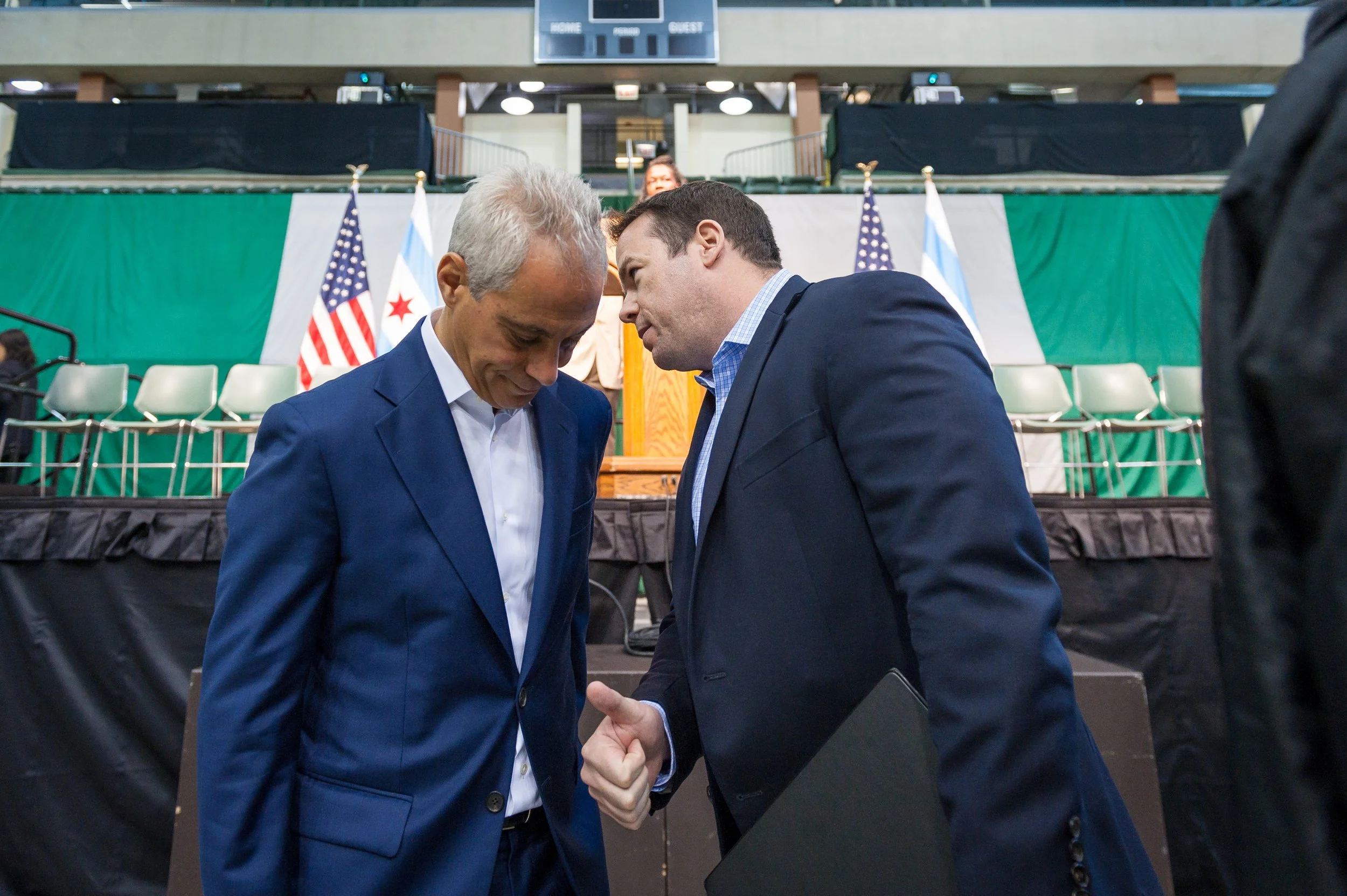 Two men in suits, one silver-haired and the other dark-haired, greeting each other with a fist bump in front of a stage with American and Illinois flags, green and white background, and empty chairs.
