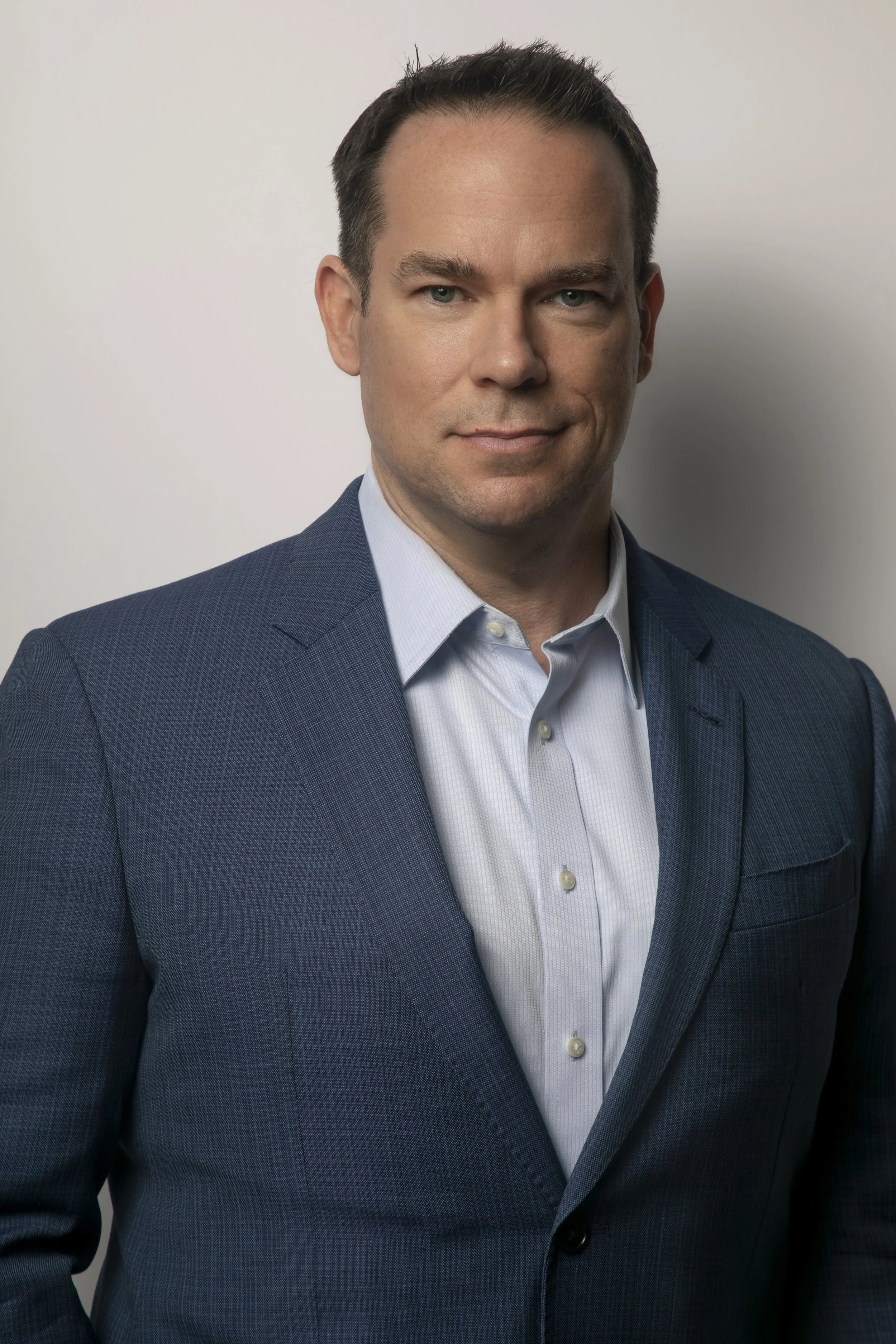 A man wearing a blue blazer and a white shirt, standing against a plain background.