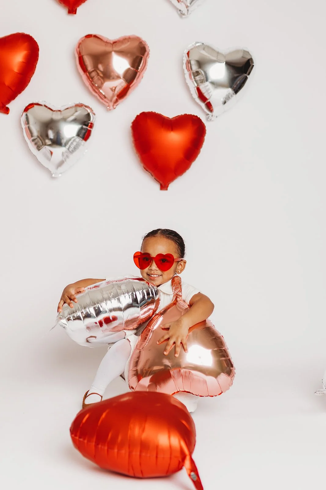 A young girl wearing red heart-shaped sunglasses sitting on the floor surrounded by red and silver heart-shaped balloons, with some balloons floating on the white background.