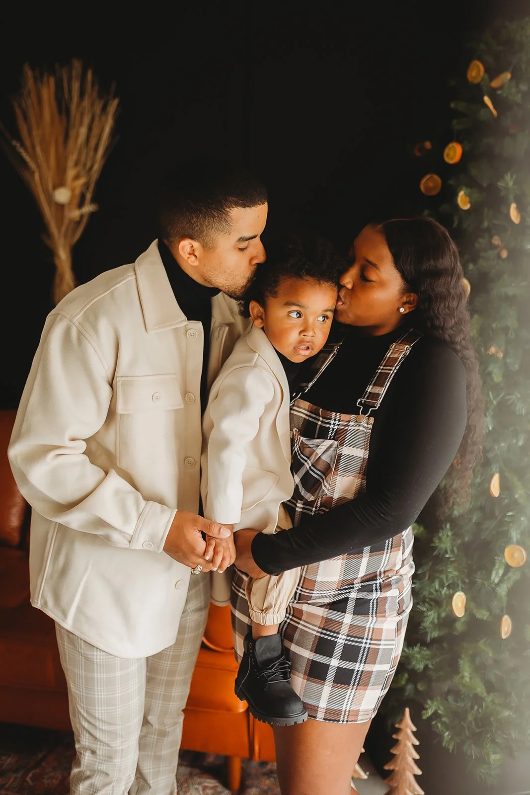 A family of three, a man, a woman, and a young girl, sharing a kiss during a holiday celebration with a decorated Christmas tree in the background.
