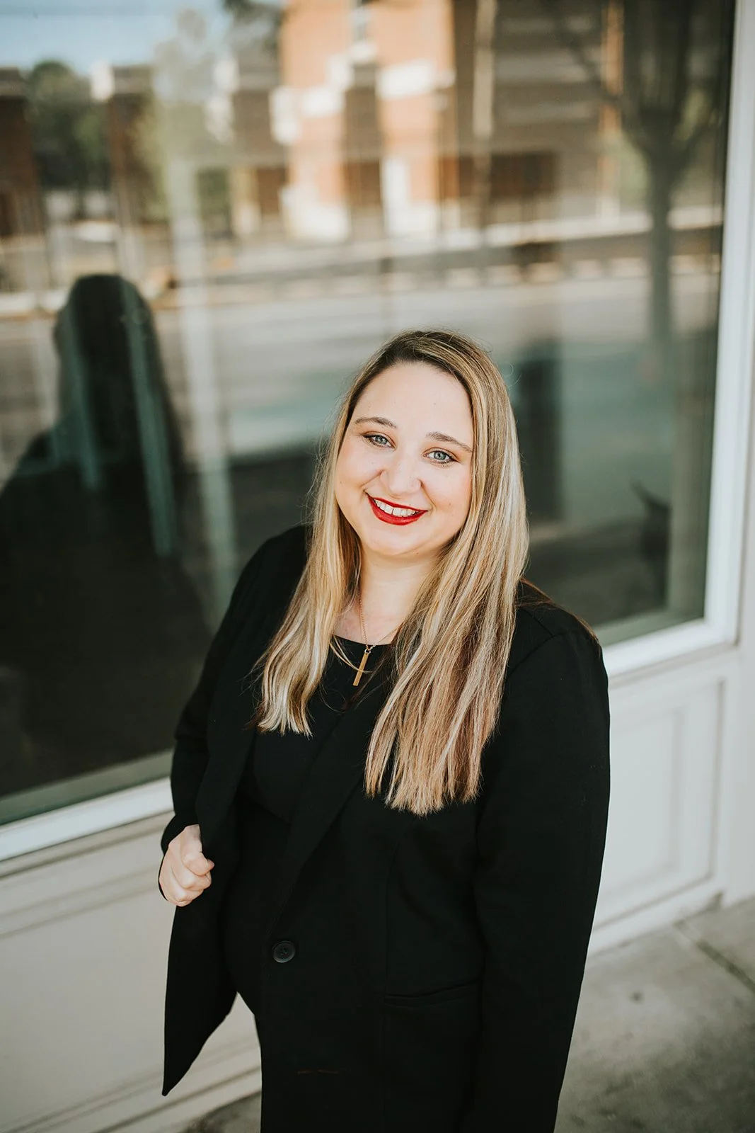 A woman with long blonde hair, wearing a black blazer and red lipstick, standing indoors in front of a large window, smiling at the camera.