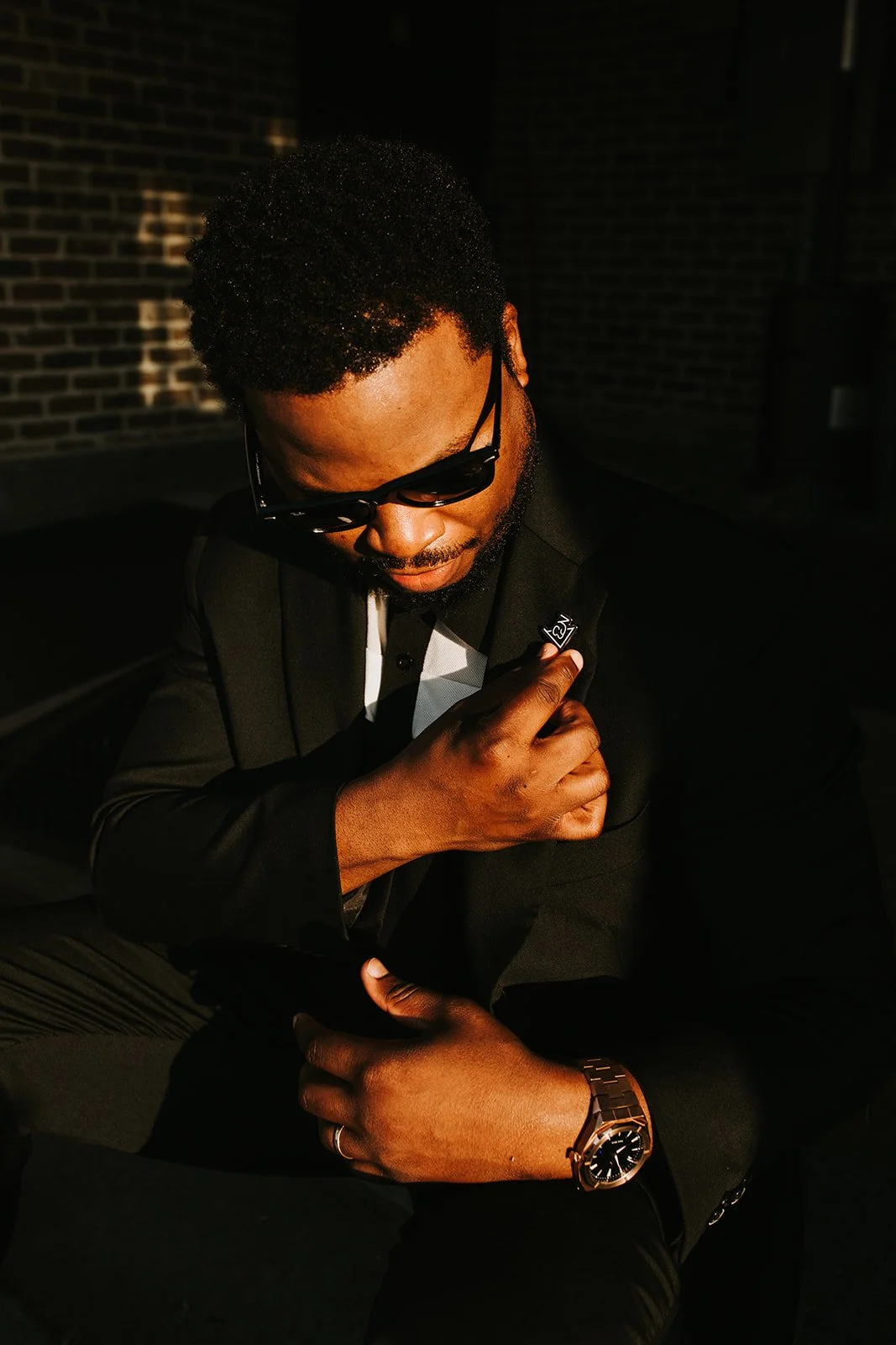 A man wearing sunglasses, a black suit, and a watch, sitting against a dark background with a brick wall, looking down and adjusting his blazer.
