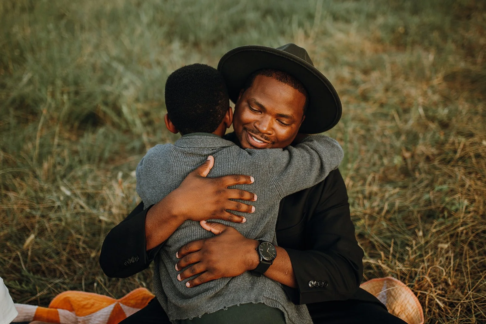 A man wearing a black hat and black jacket hugging a young boy dressed in gray, both smiling with eyes closed, sitting on the grass in an outdoor setting.