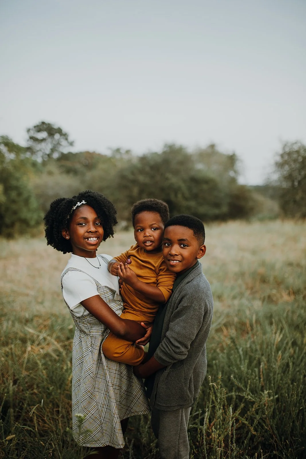 Three children standing outdoors in a field with trees in the background, smiling and looking at the camera, with the youngest being held by the girl on the left.