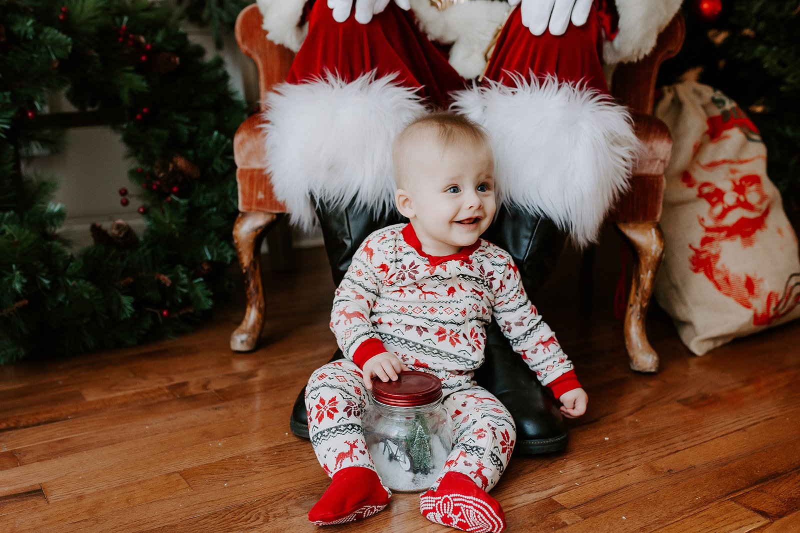 A young child in Christmas pajamas sitting on a wooden floor, holding a jar with a small Christmas tree inside, smiling happily. Behind him, a Santa Claus figure is seated in a decorated Christmas setting with a Christmas tree and festive decorations.