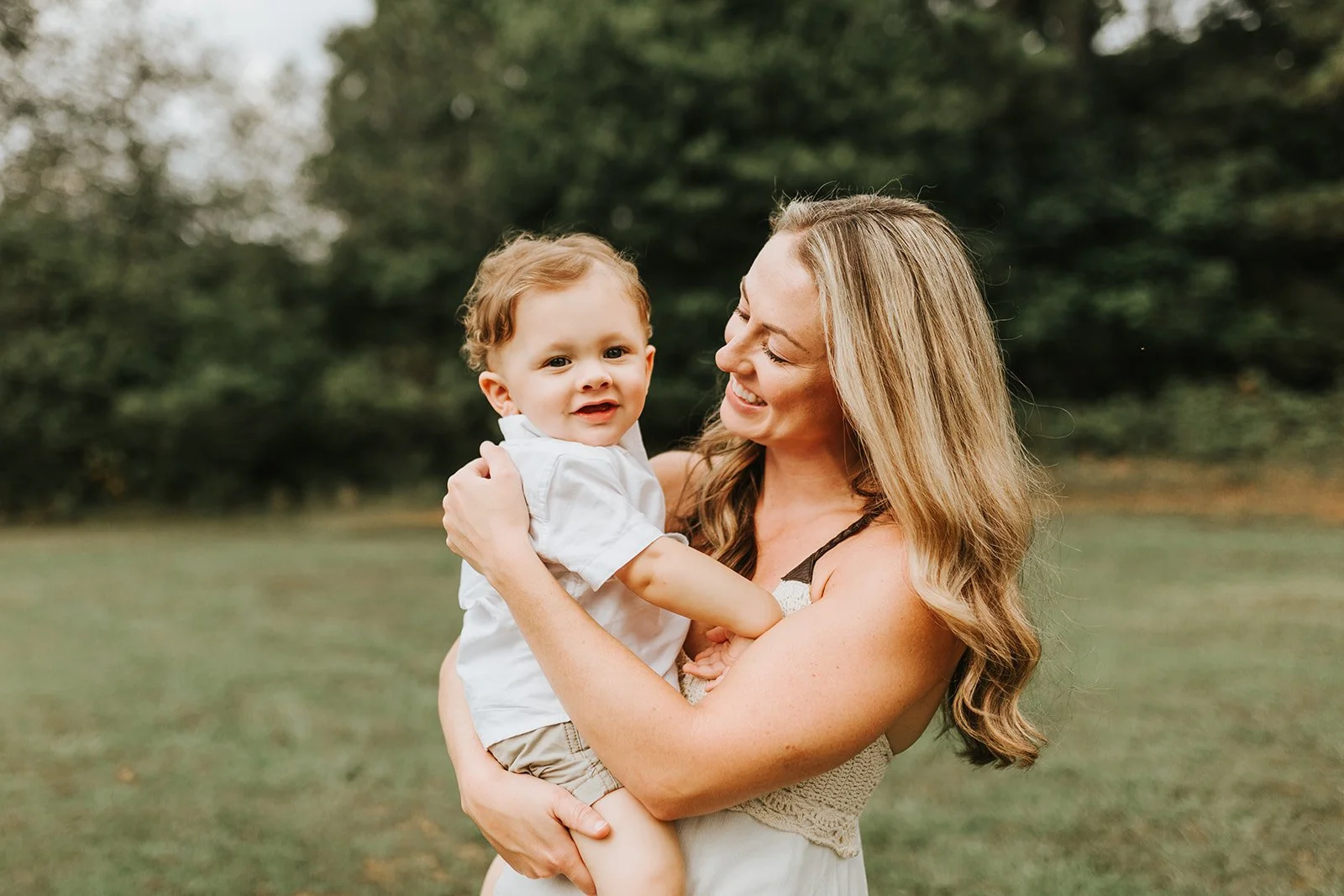 A woman holding a young boy outdoors in a grassy area with trees in the background, smiling and looking at each other.
