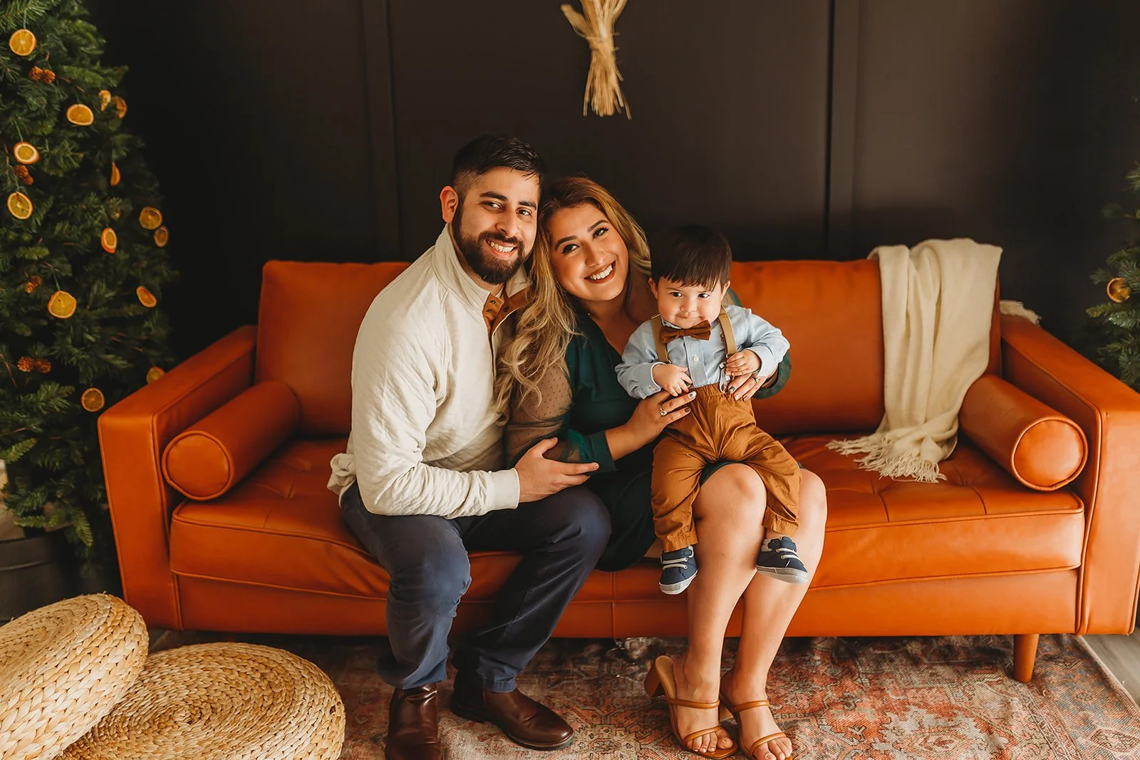 A family of three, a father, mother, and young son, smiling and sitting on an orange sofa. The background includes Christmas trees decorated with orange slices. The family appears happy, with the mother and son close together, and the father beside them. There are woven baskets on the floor.