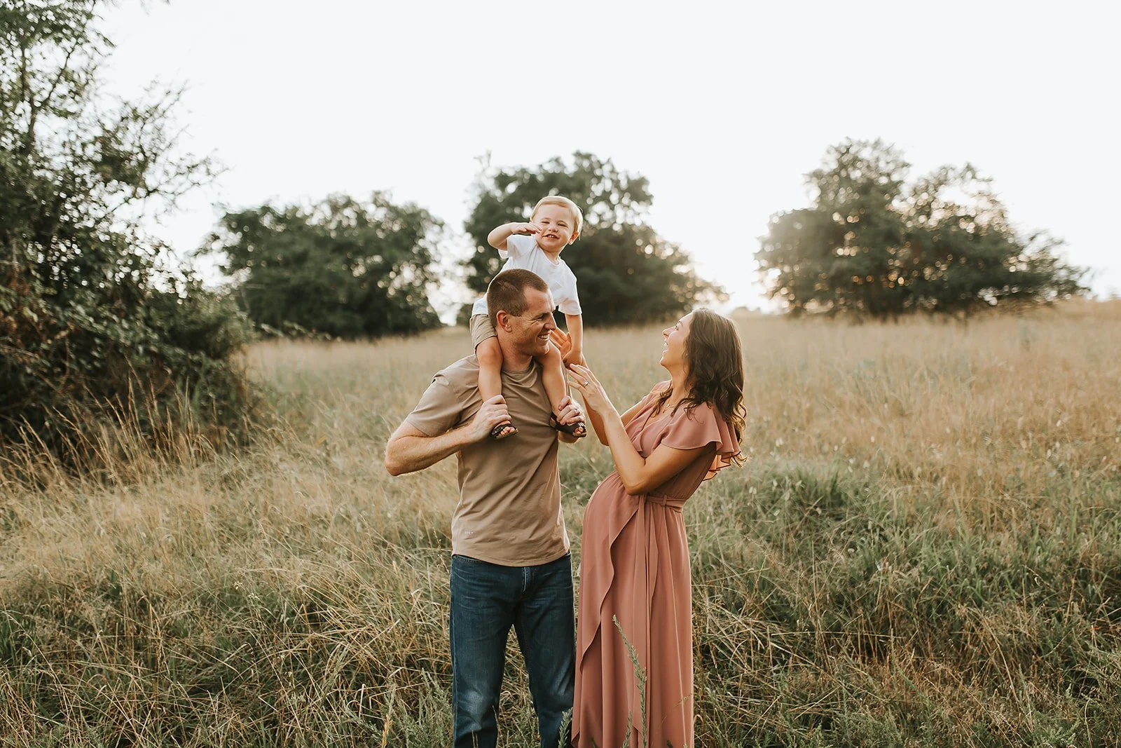 A family of three enjoying an outdoor walk in a grassy field during sunset. The father carries the young son on his shoulders, and the mother gently touches the son's foot, smiling at each other.